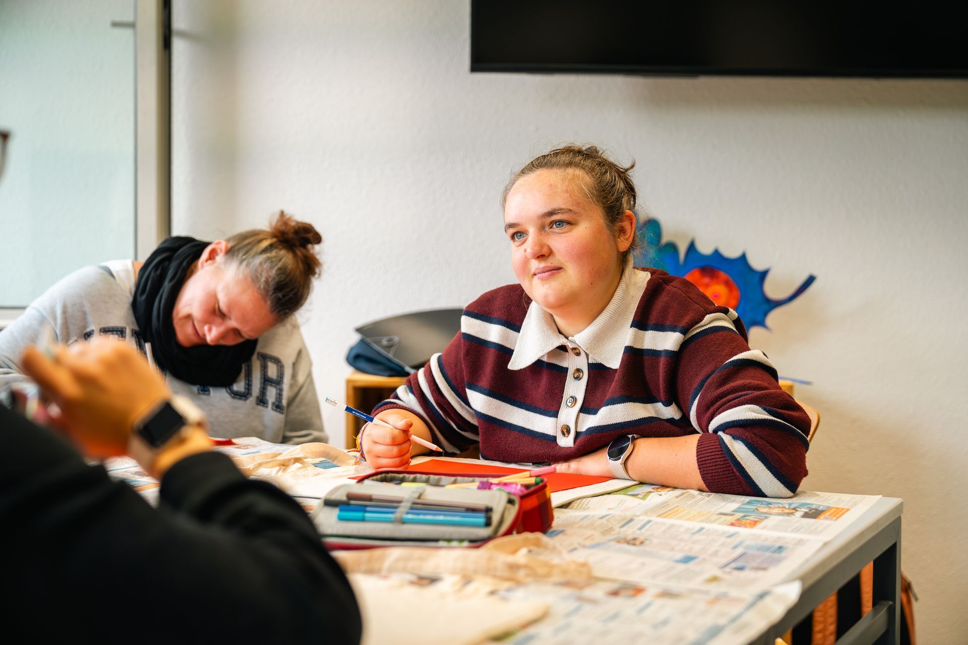 Eine Frau in einem gestreiften Hemd lächelt, sie sitzt mit anderen an einem Tisch; Malutensilien sind zu sehen. Eine weitere Person lehnt sich daneben.