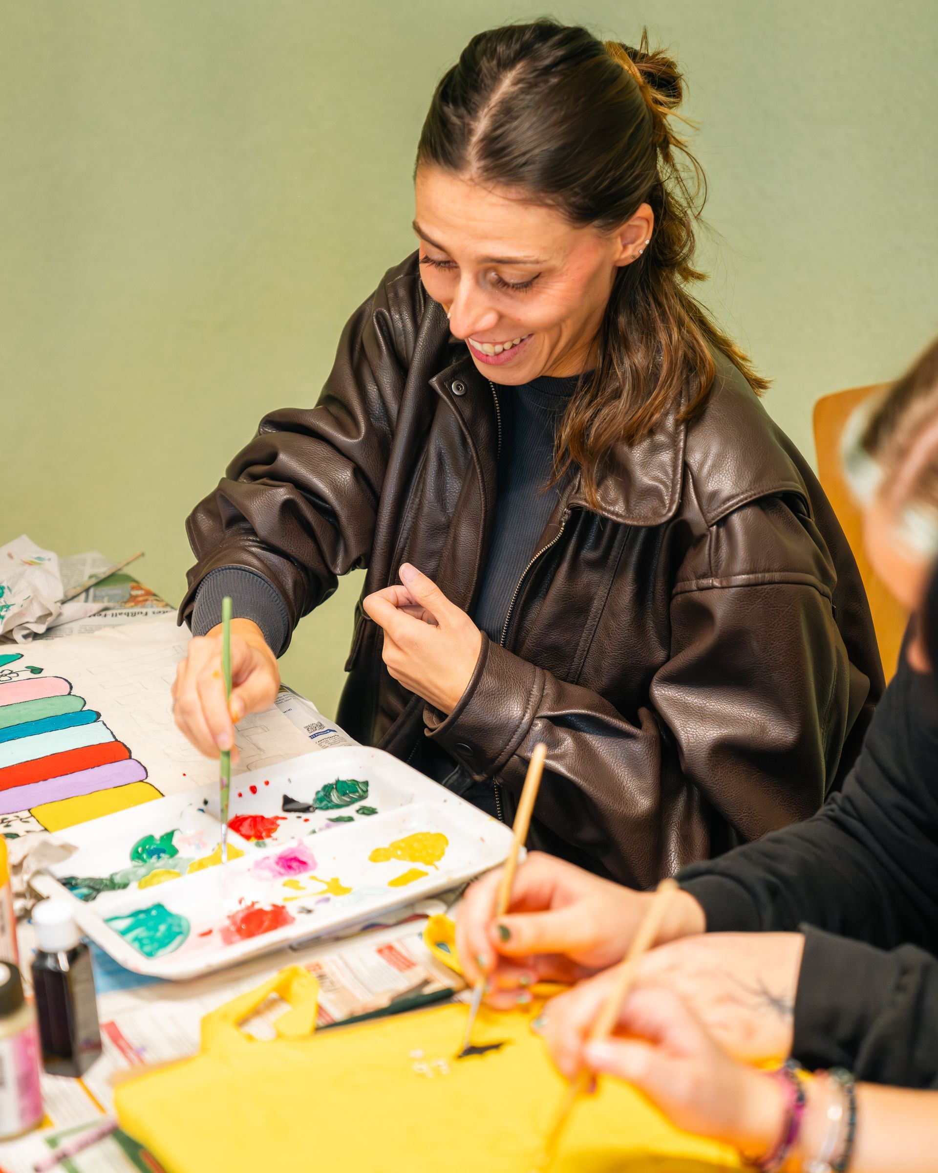 Eine Frau in brauner Jacke malt an einem Tisch mit einer Palette. Sie lächelt, während um sie herum andere malen.