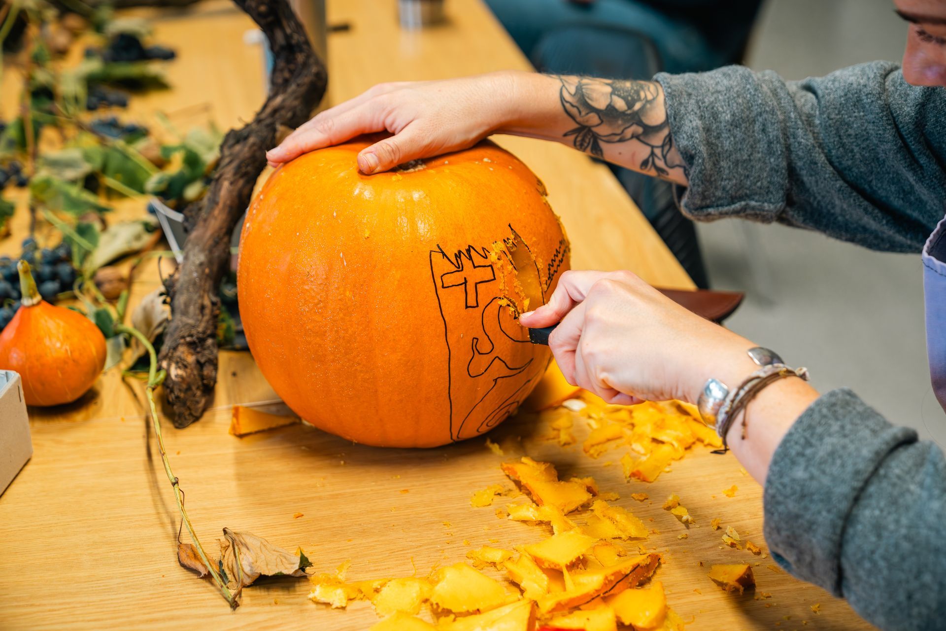 Eine Person schnitzt mit einem Messer einen Kürbis. Der Kürbis ist orange. Die Schnitzerei zeigt eine Katze. Holztisch.