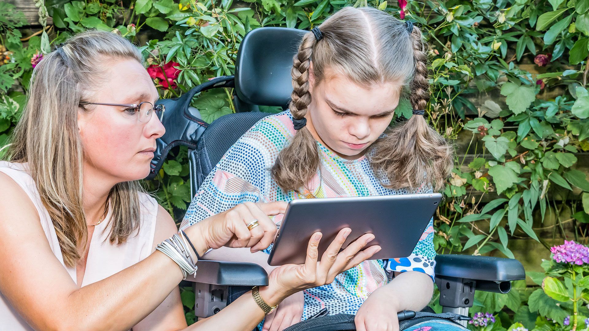 eine Frau zeigt einem Mädchen im Rollstuhl auf etwas auf dem Tablet