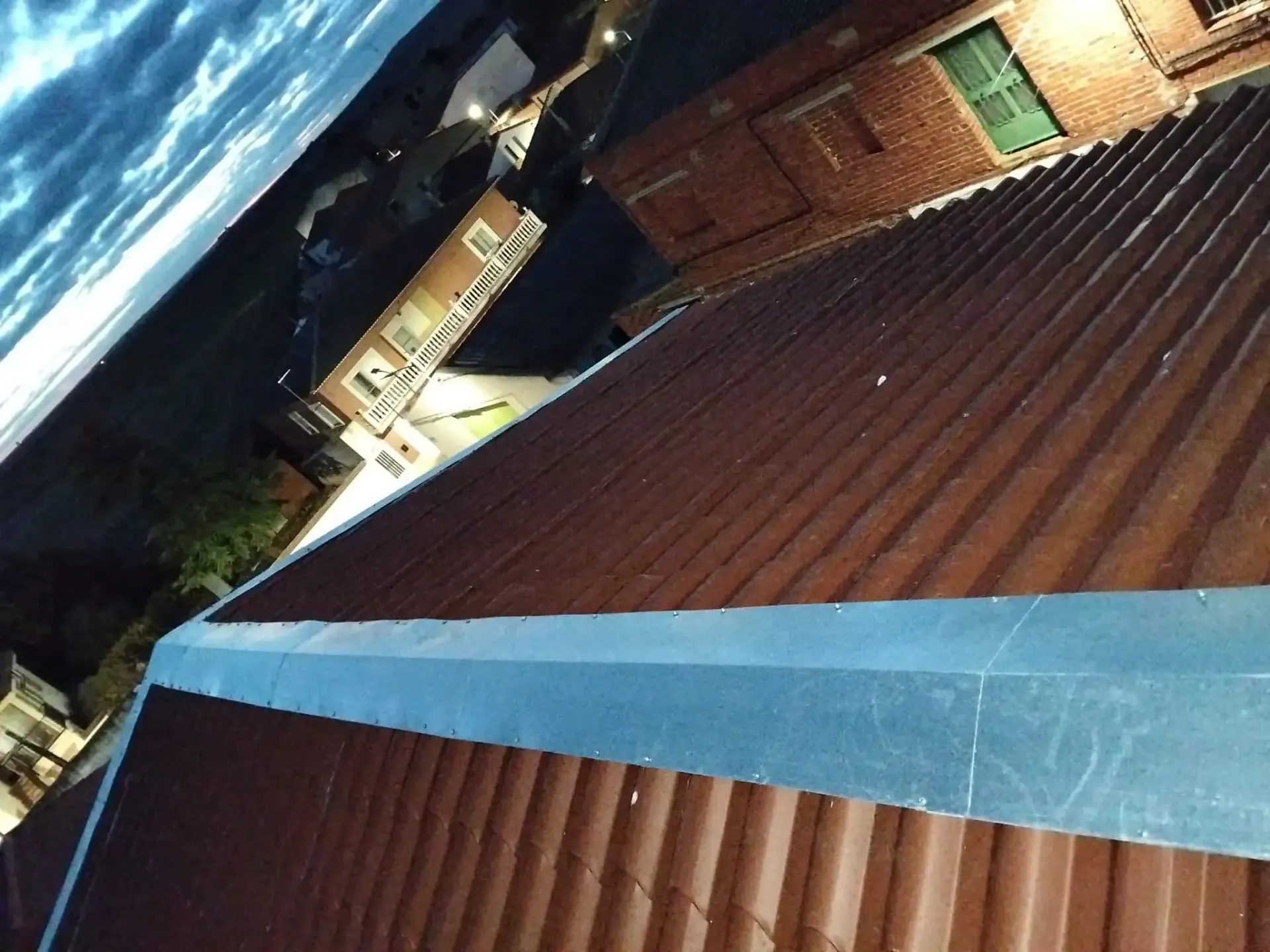 Vista aérea de un techo de metal corrugado de color marrón con borde azul, edificios y un cielo nublado.