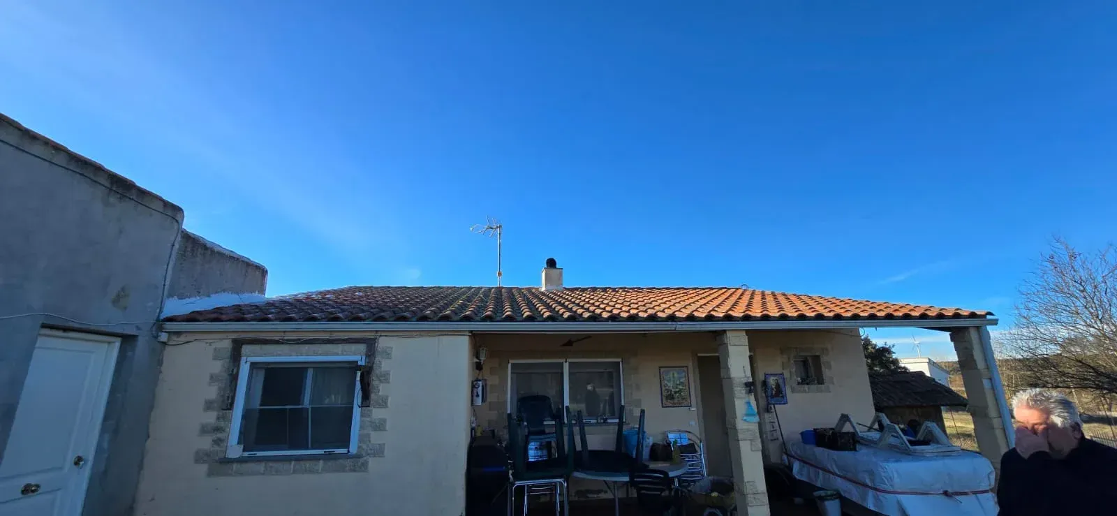 Una casa clara con techo de tejas rojas bajo un cielo azul claro. Una persona está de pie a la derecha.
