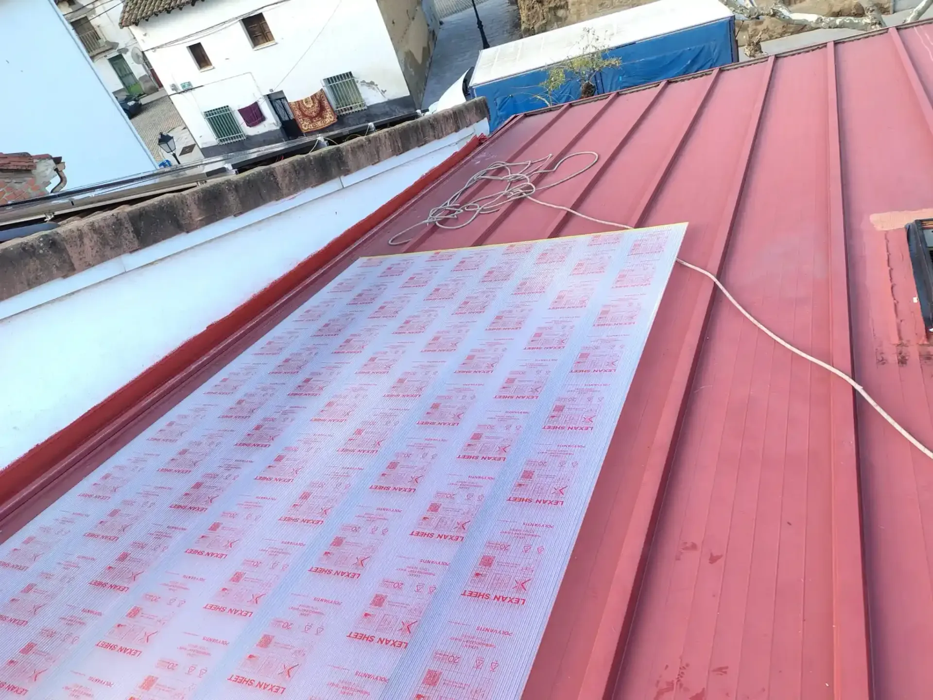 Techo de metal rojo con lámina protectora, cables y vista parcial de los edificios.