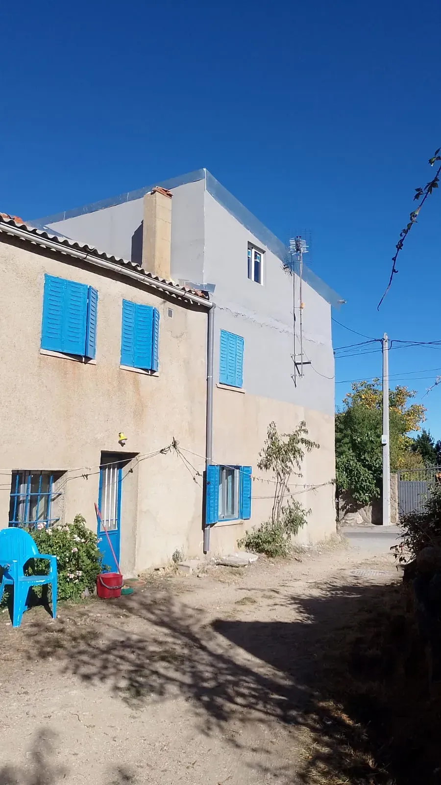 Casa de dos pisos con persianas y puerta azules; un camino de tierra pasa por delante de ella en un día soleado.