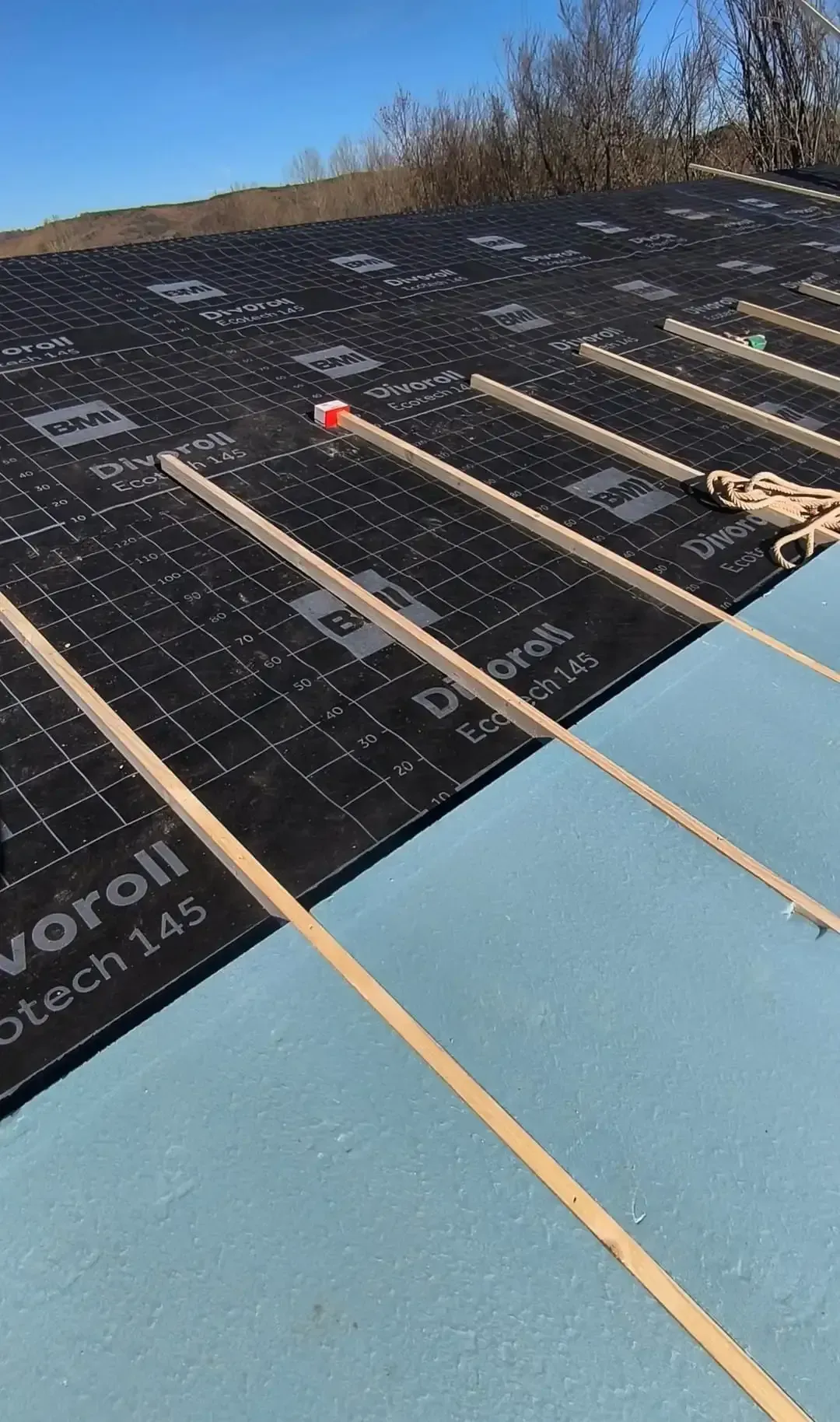 Trabajos de techado en progreso: aislamiento, capa base negra, listones de madera y un dispositivo de medición con tapa roja.