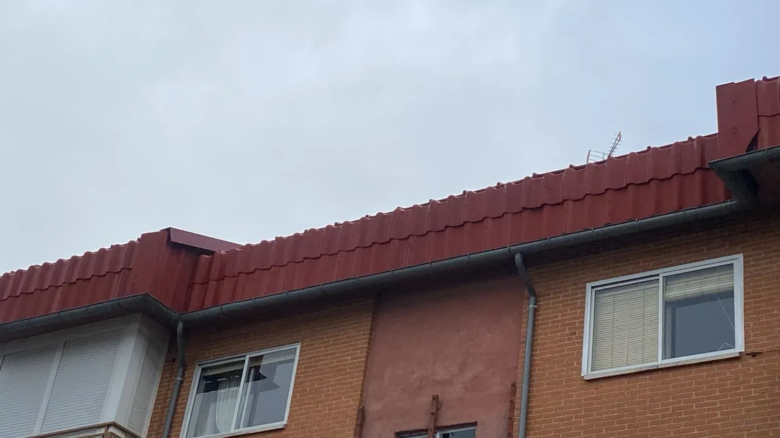 Edificio de tejado rojo con fachada de ladrillo y dos ventanas, bajo un cielo nublado.