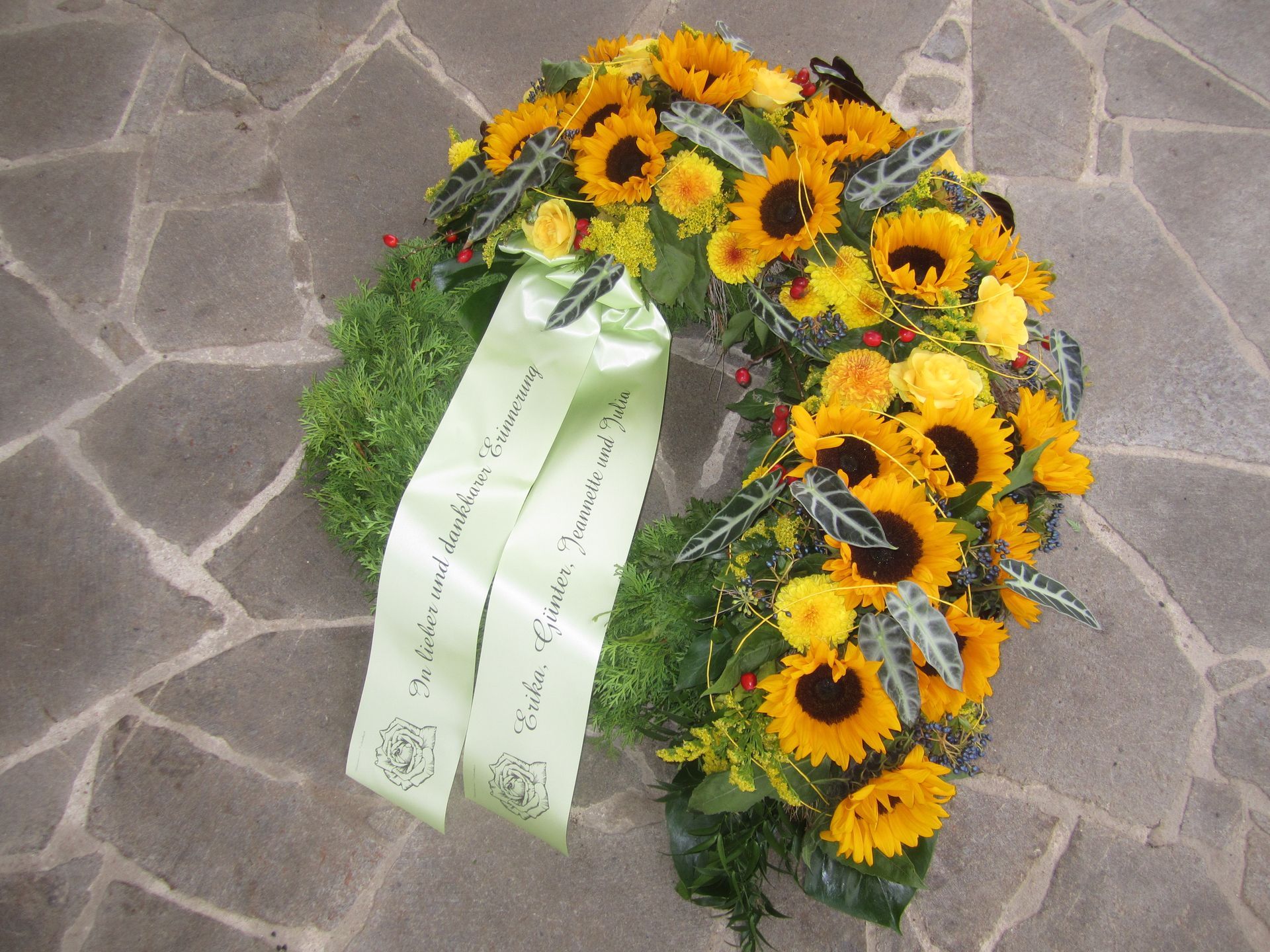 Kranz aus Sonnenblumen und Grünpflanzen mit einem grünen Band auf einer Steinoberfläche.