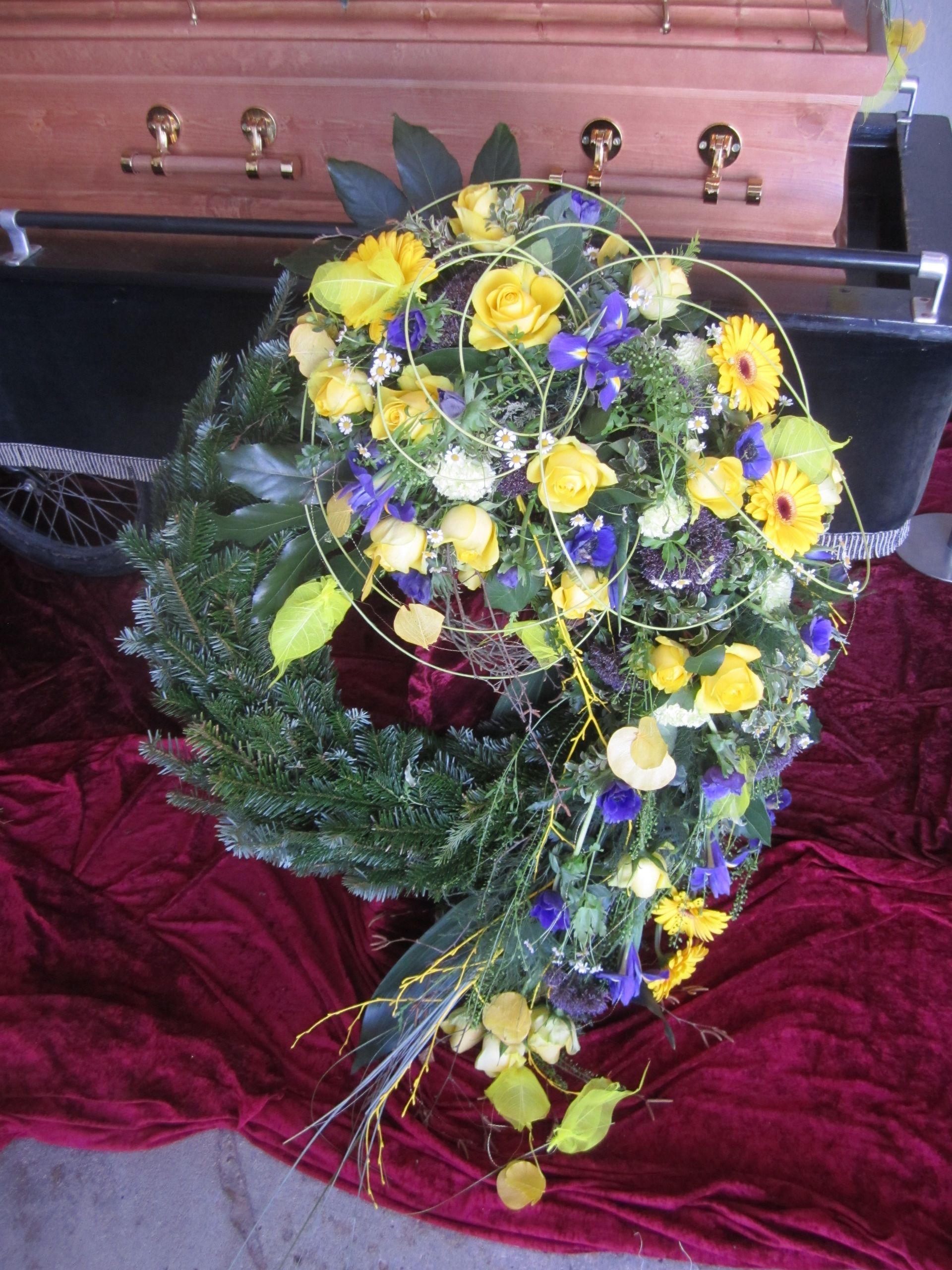 Trauerkranz mit gelben, weißen und violetten Blumen, roter Stoffhintergrund, im Hintergrund ein Sarg.