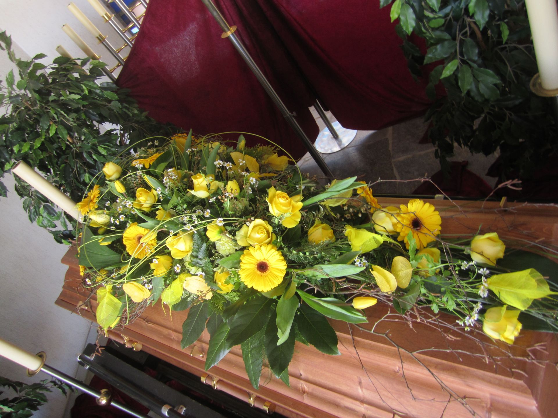 Gelbes Blumenarrangement auf einem Holzsarg bei einer Beerdigung; roter Hintergrund und Grün.