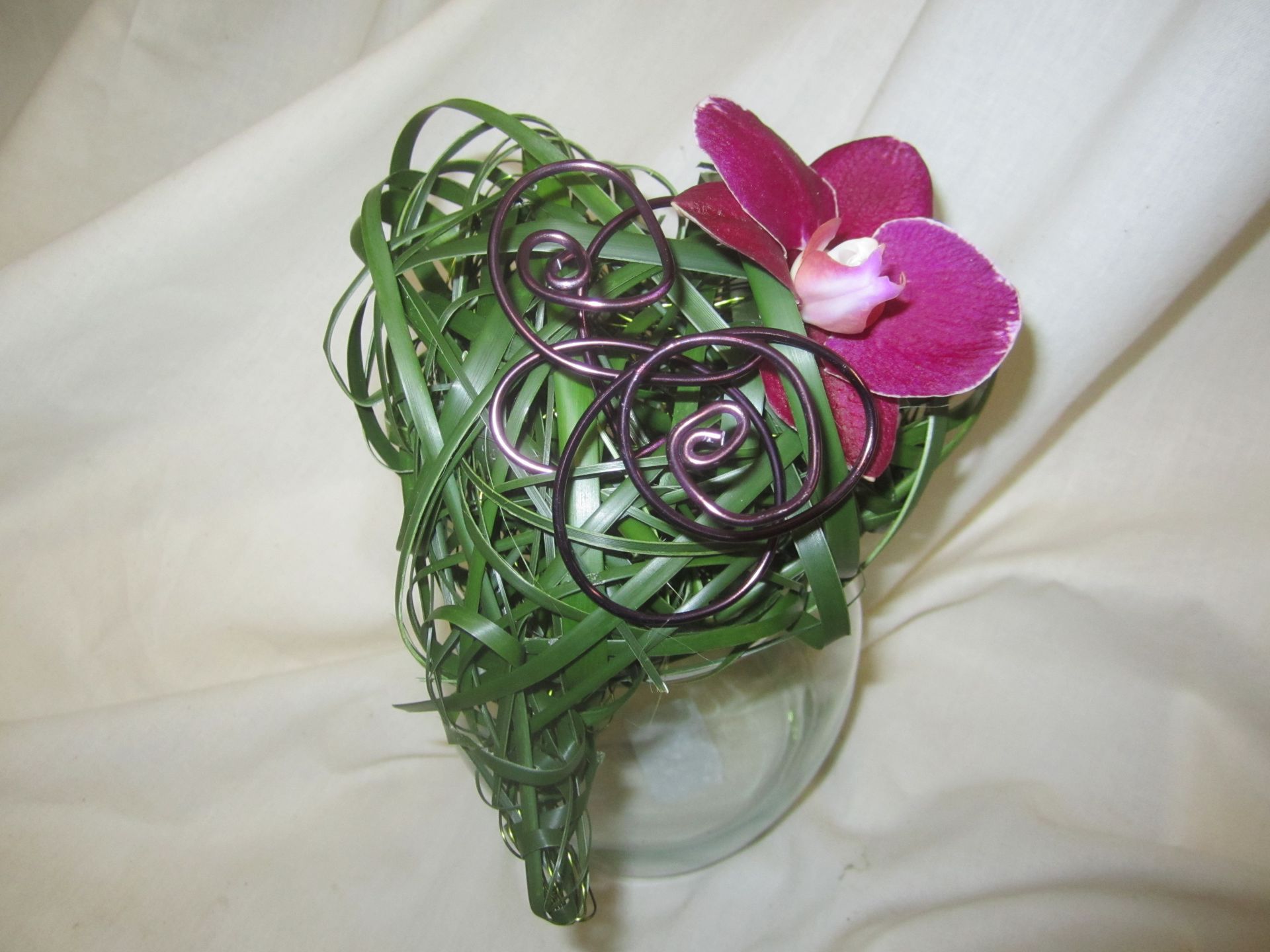 Grünes Blumenarrangement mit lila Orchidee und Metallwirbeln auf einer weißen Stoffserviette.