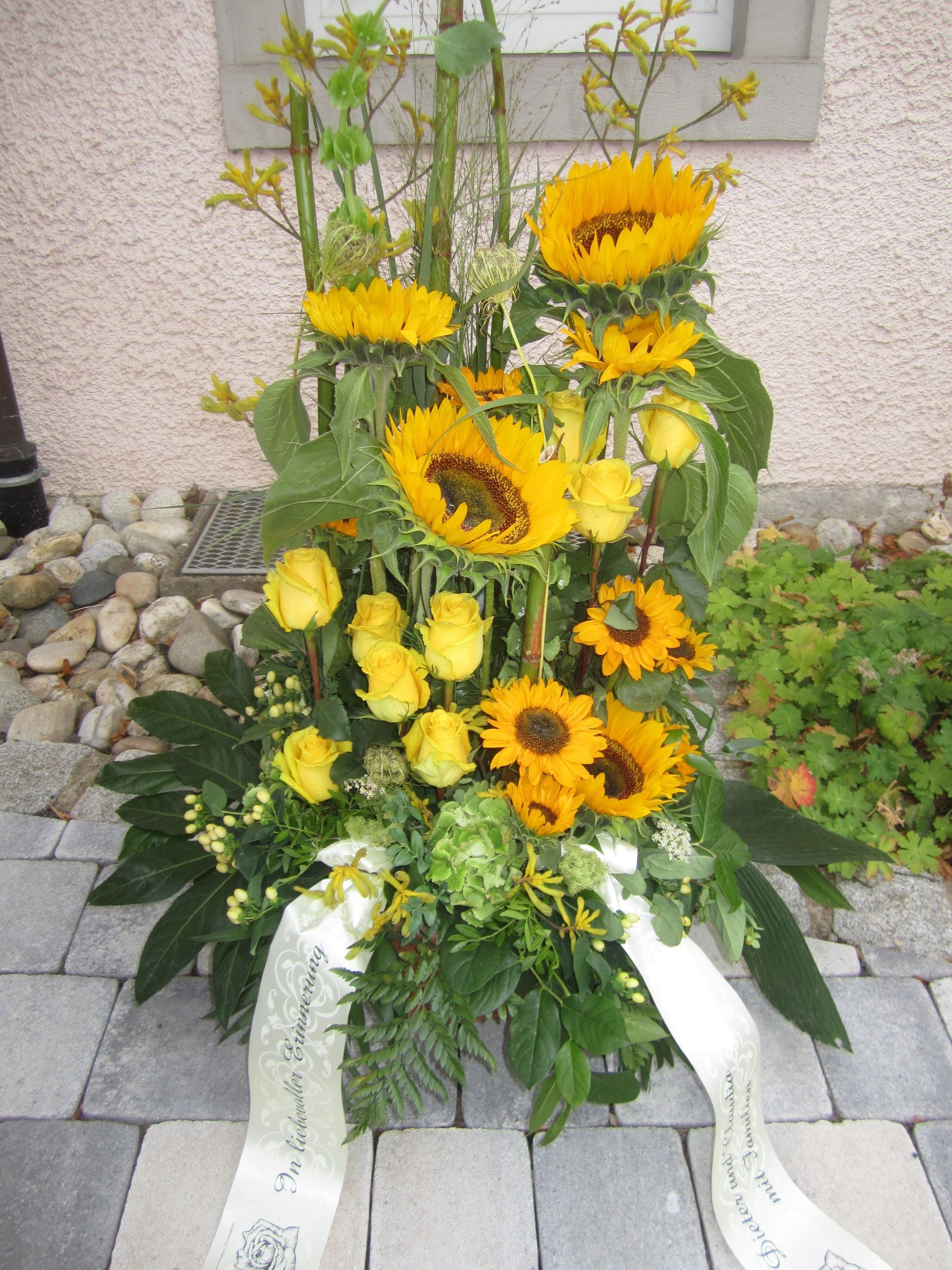 Gelbes Blumenarrangement mit Sonnenblumen, Rosen und Bändern, im Freien auf Steinpflaster.