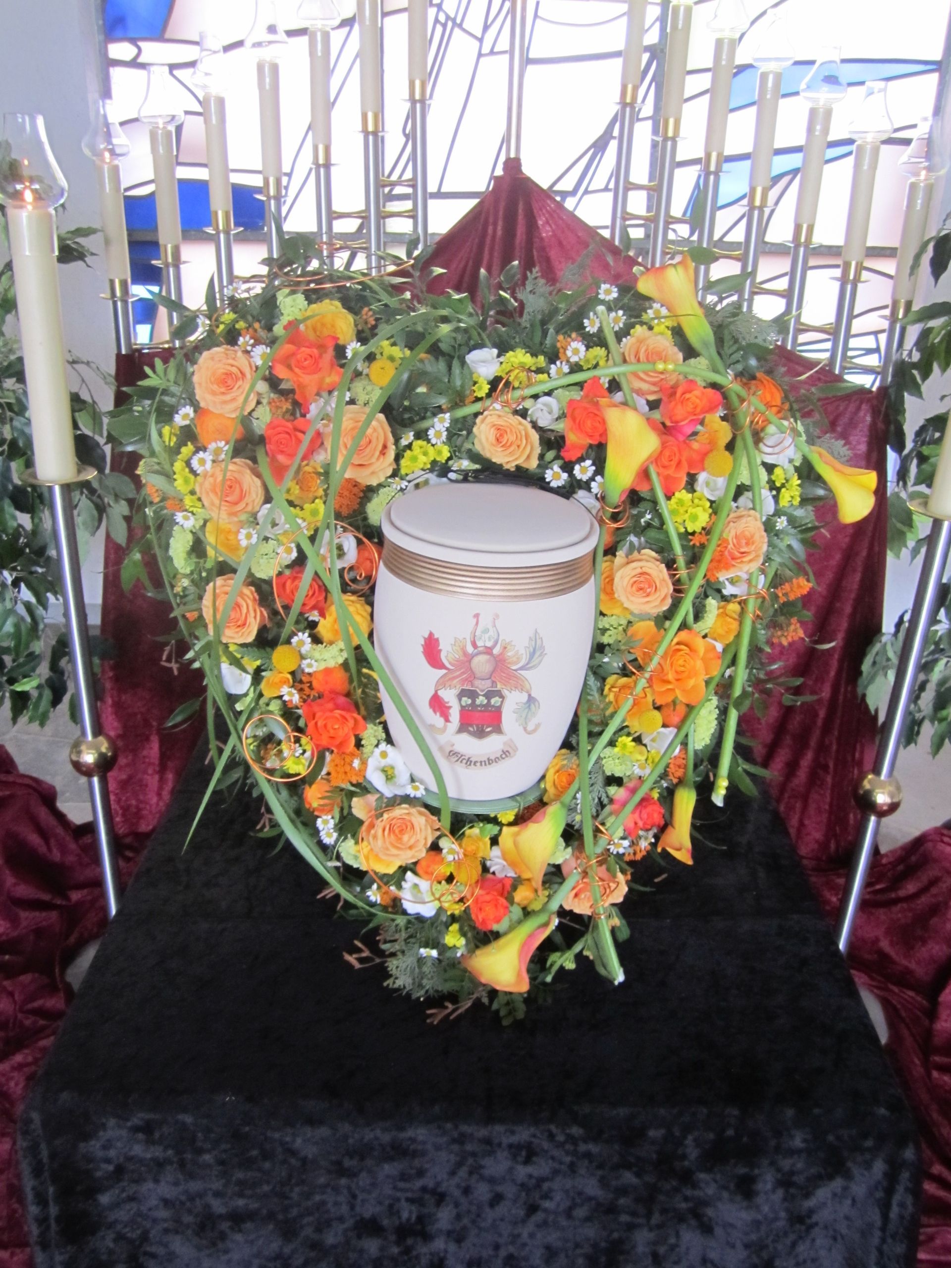 Herzförmiges Blumenarrangement mit Urne bei einer Trauerfeier. Orange und gelbe Blumen, burgunderfarbener Hintergrund, Kerzen in einer kirchlichen Umgebung.