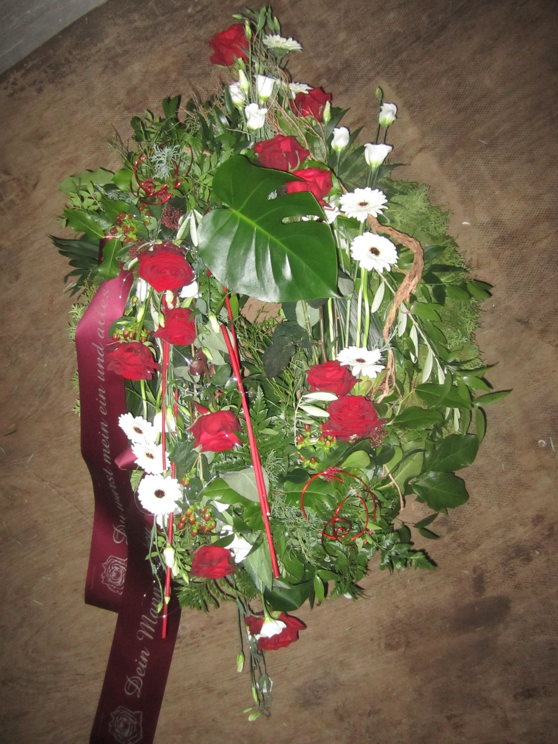 Kranz aus roten Rosen, weißen Blumen, grünem Laub und Band vor braunem Hintergrund.