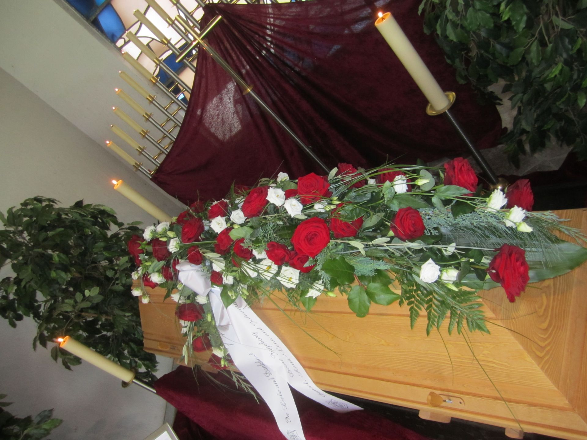 Sarg mit Blumen und weißem Band geschmückt; Kerzen und burgunderfarbener Hintergrund im Trauerumfeld.