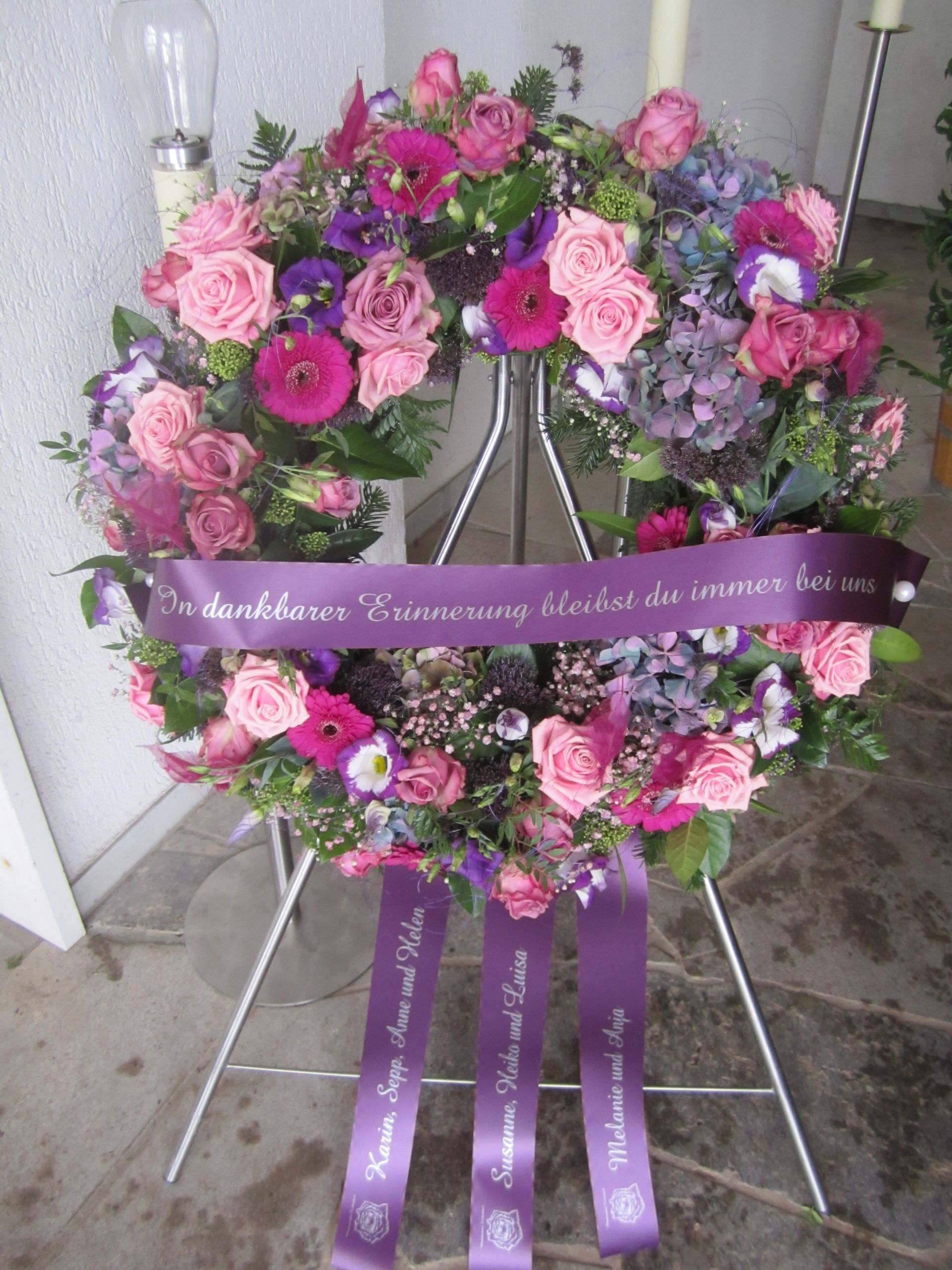 Ein Blumenkranz in Rosa- und Lilatönen, präsentiert auf einem Metallständer mit einem lila Band.