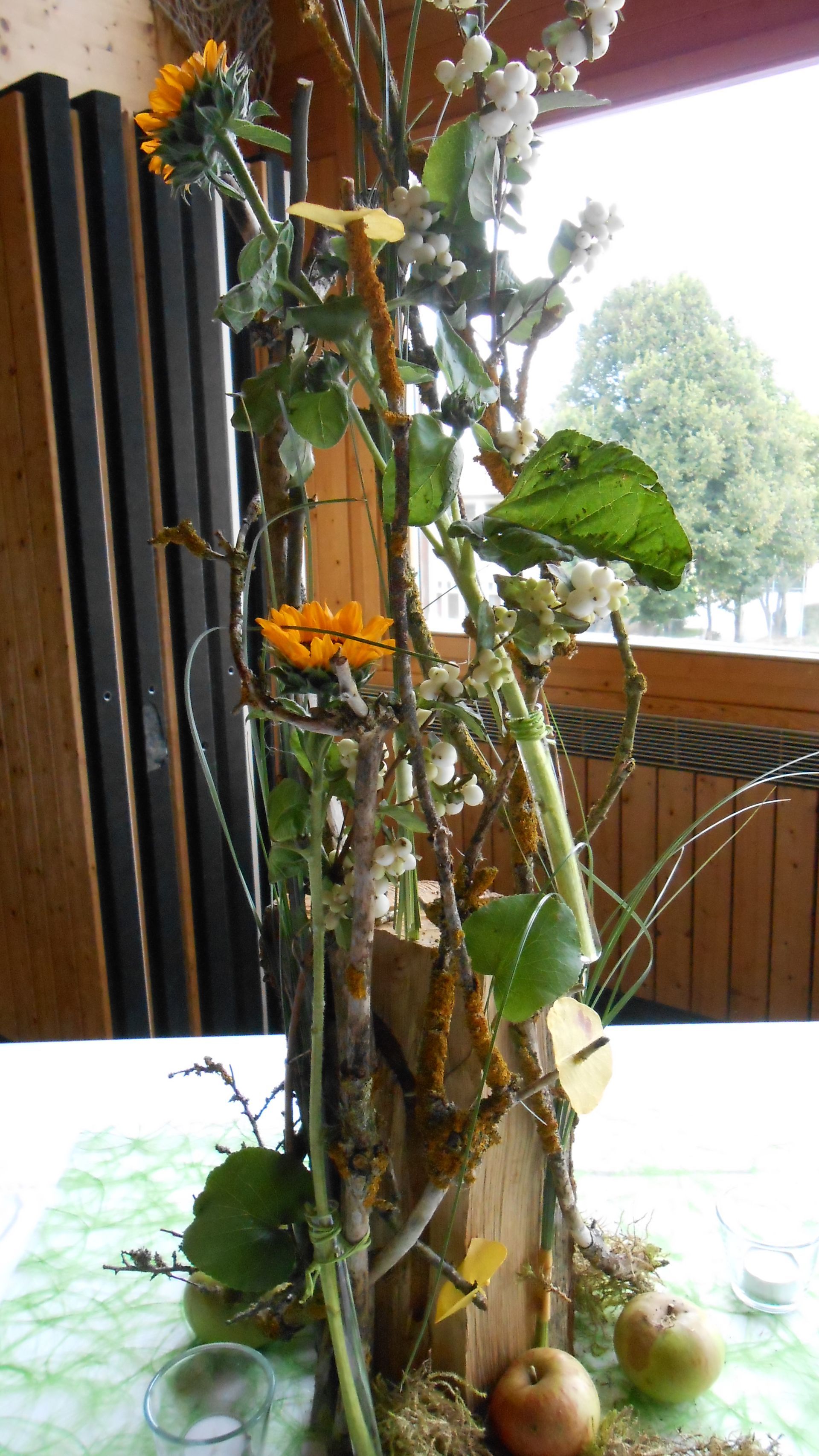 Tafelaufsatz mit Sonnenblumen, Äpfeln und Zweigen auf einem Tisch mit grüner Tischdecke.