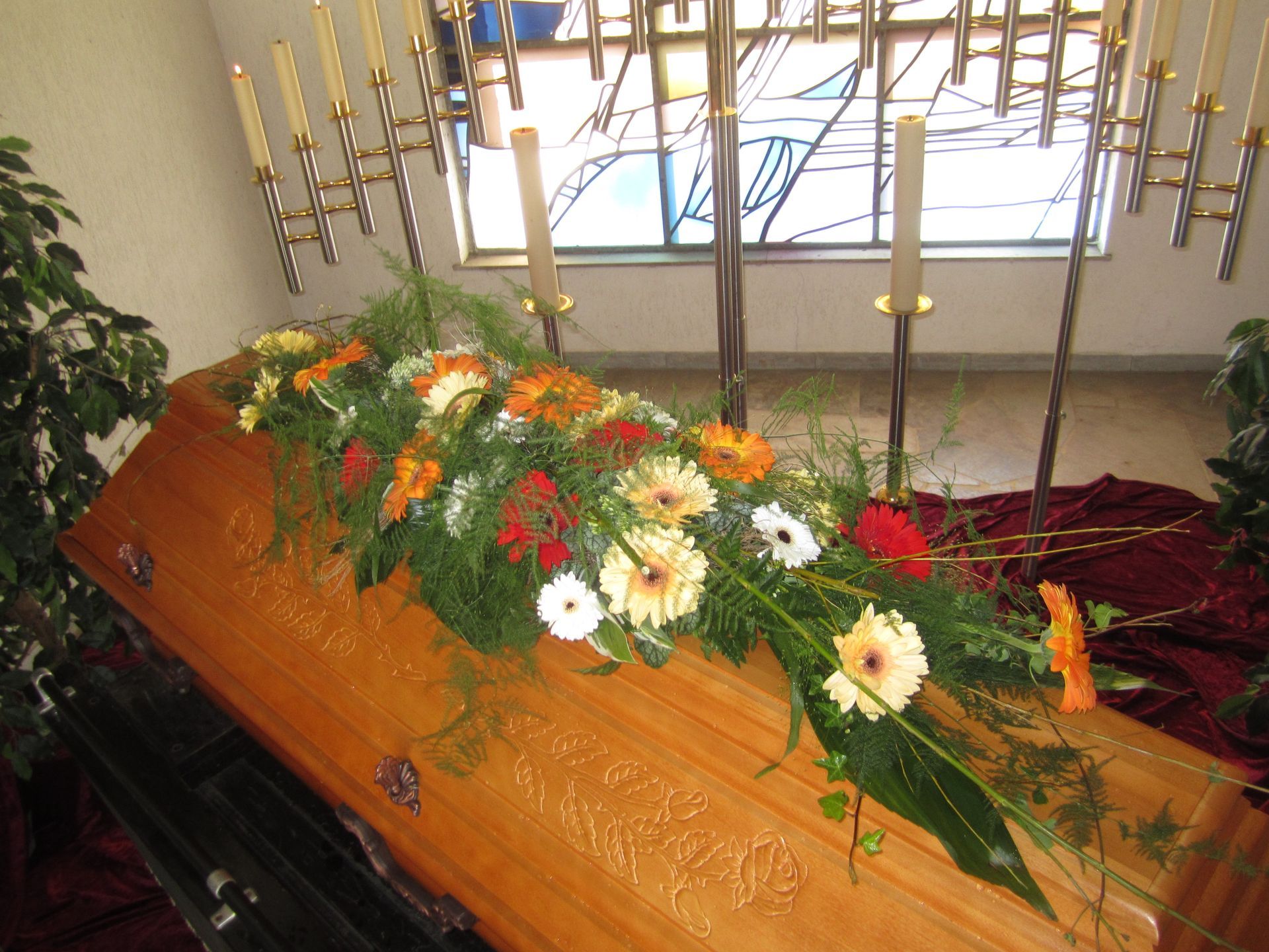 Sarg mit Blumenarrangement bei einer Beerdigung. Gerbera-Gänseblümchen, Grünpflanzen, Kerzen im Hintergrund.