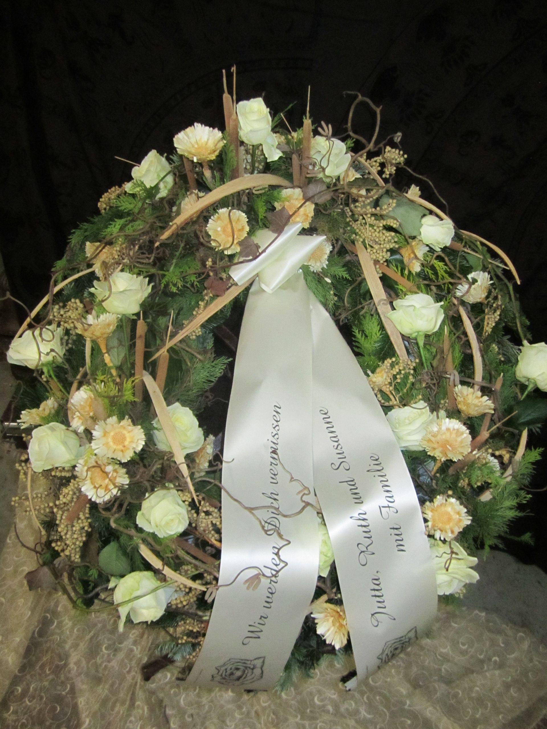 Kranz aus weißen Rosen und Nelken mit Bandinschrift; auf einer Steinoberfläche.
