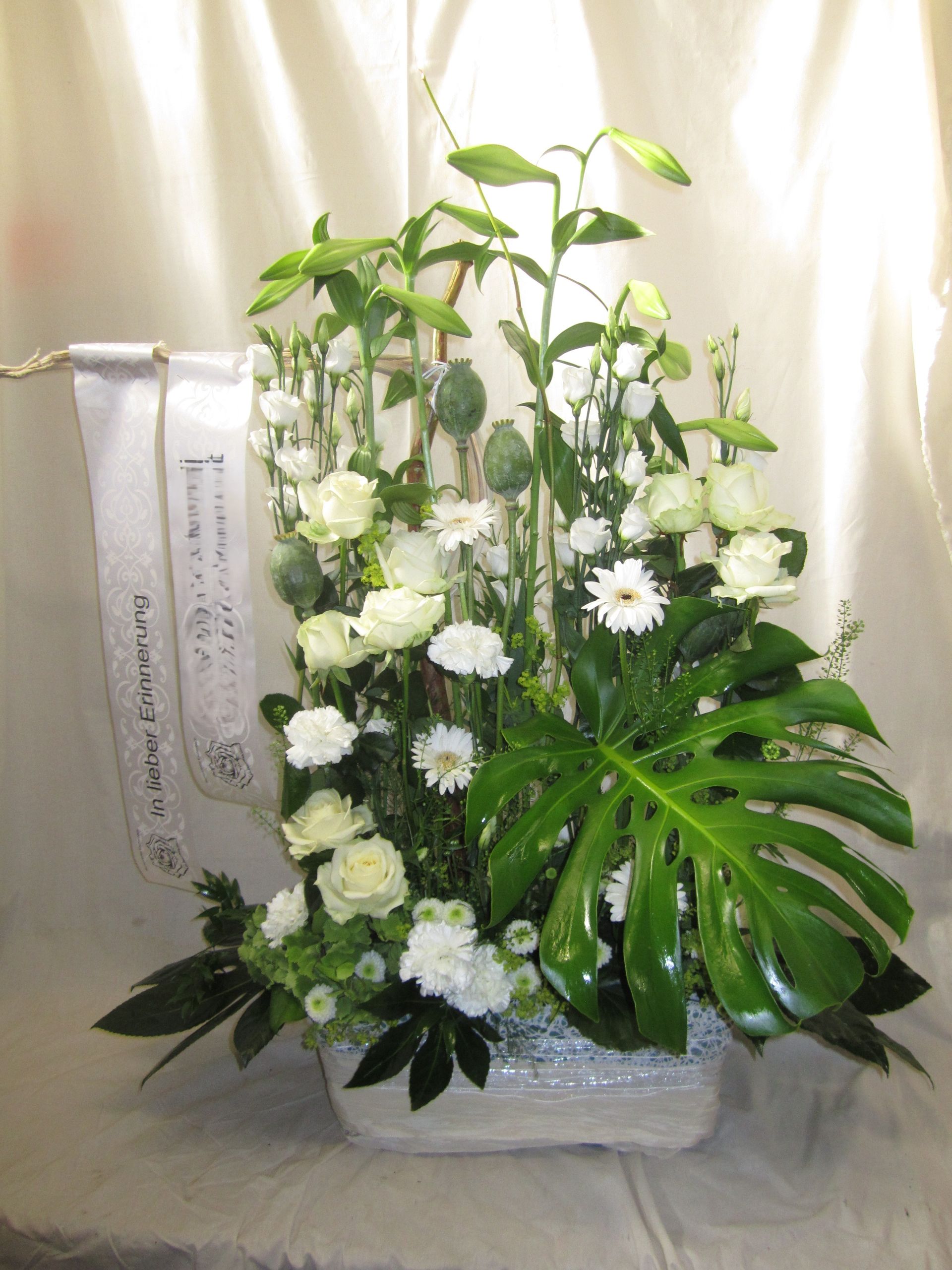 Weißes Blumenarrangement in einem weißen rechteckigen Korb mit einem grünen Monstera-Blatt und einem weißen Band.