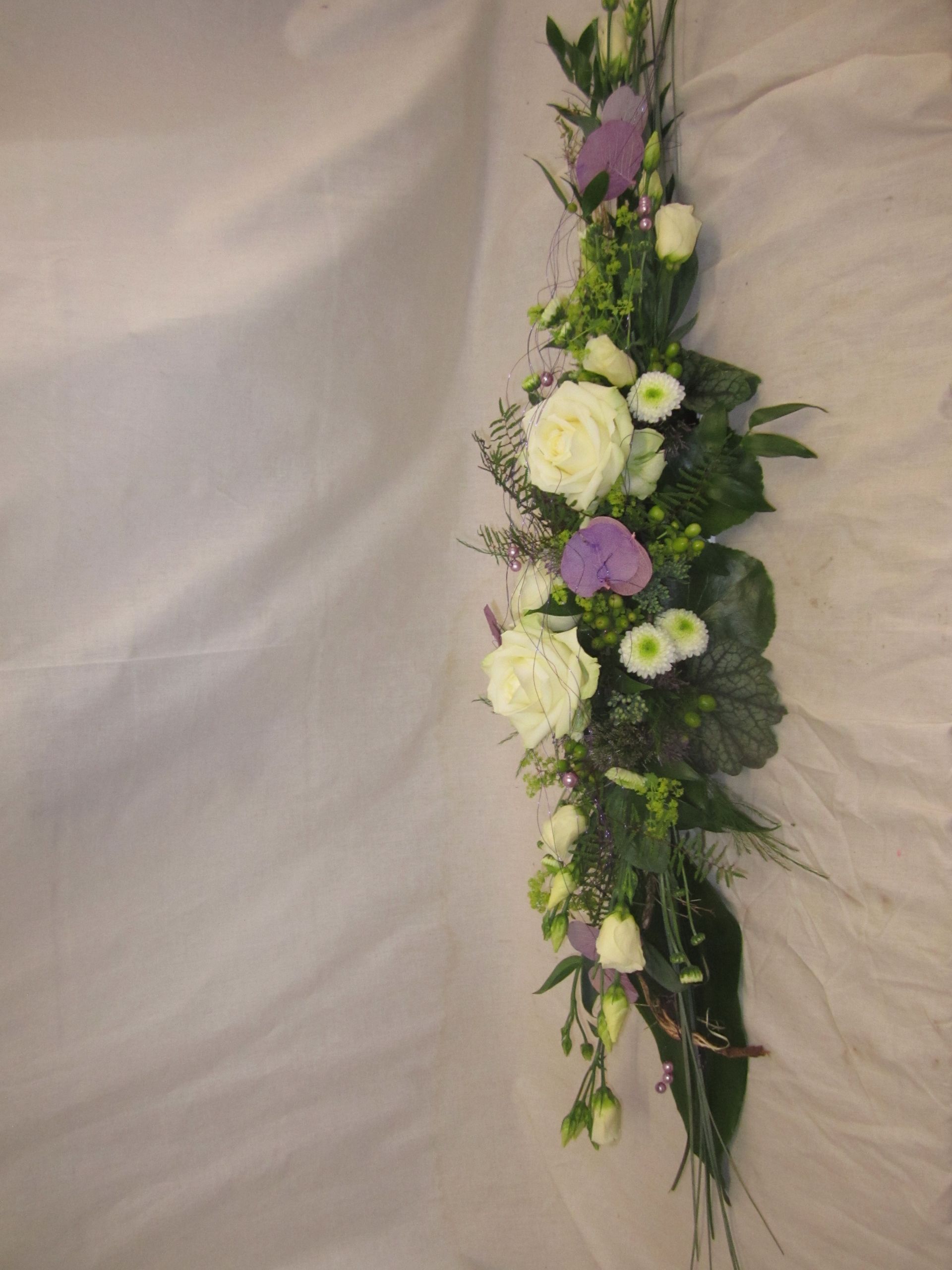 Vertikales Blumenarrangement aus weißen Rosen, violetten Akzenten und Grünpflanzen vor einem weißen Hintergrund.