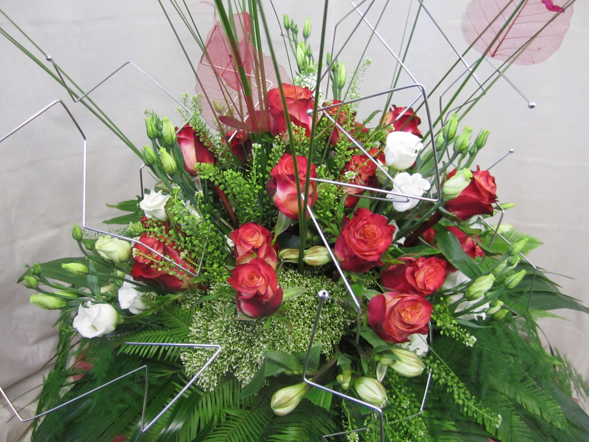 Blumenarrangement mit roten Rosen, weißen Blumen und geometrischen Drahtakzenten.