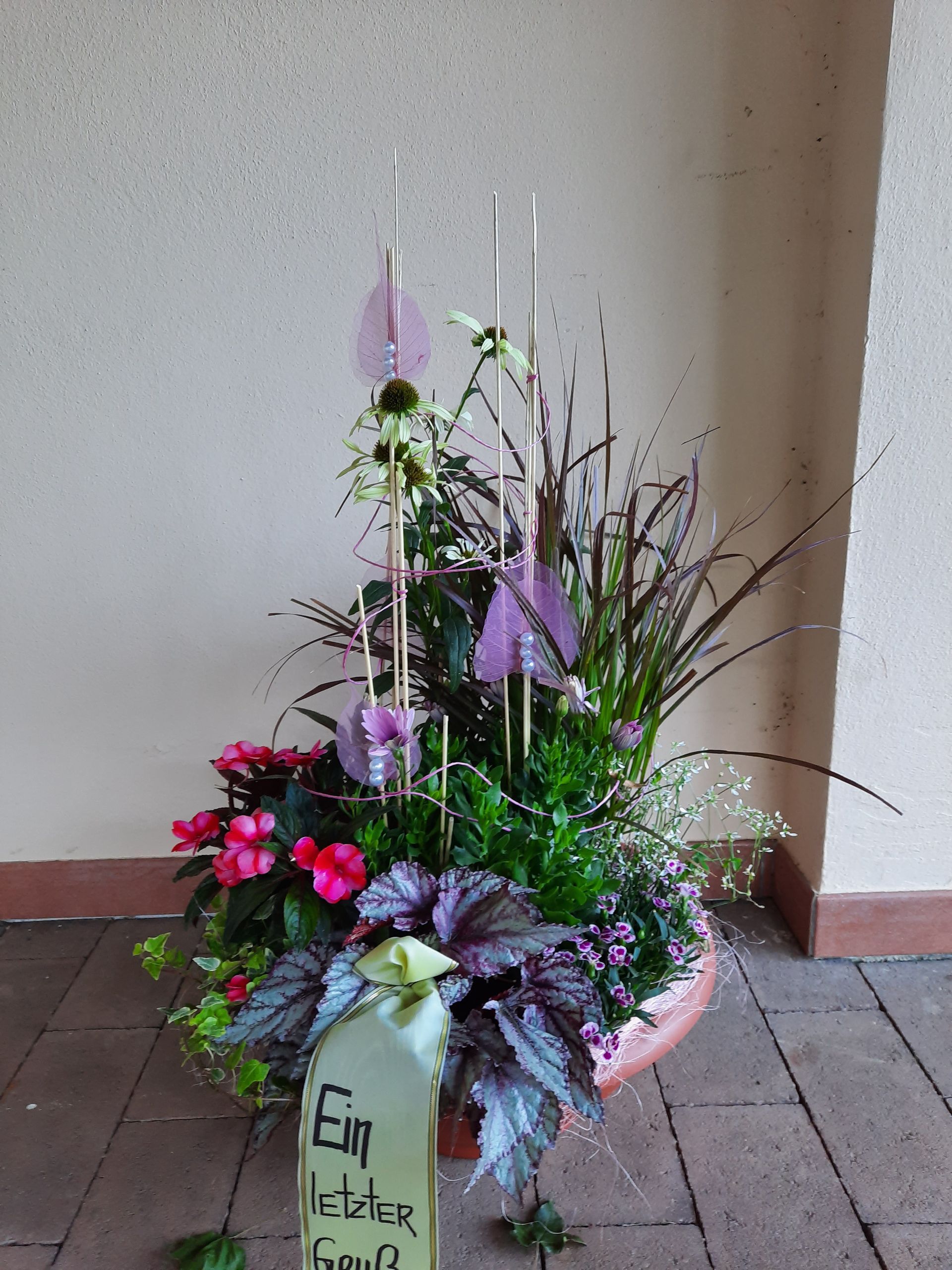 Blumenarrangement mit rosa Blumen, Grünpflanzen, Metallstäben und einem Gedenkschild.