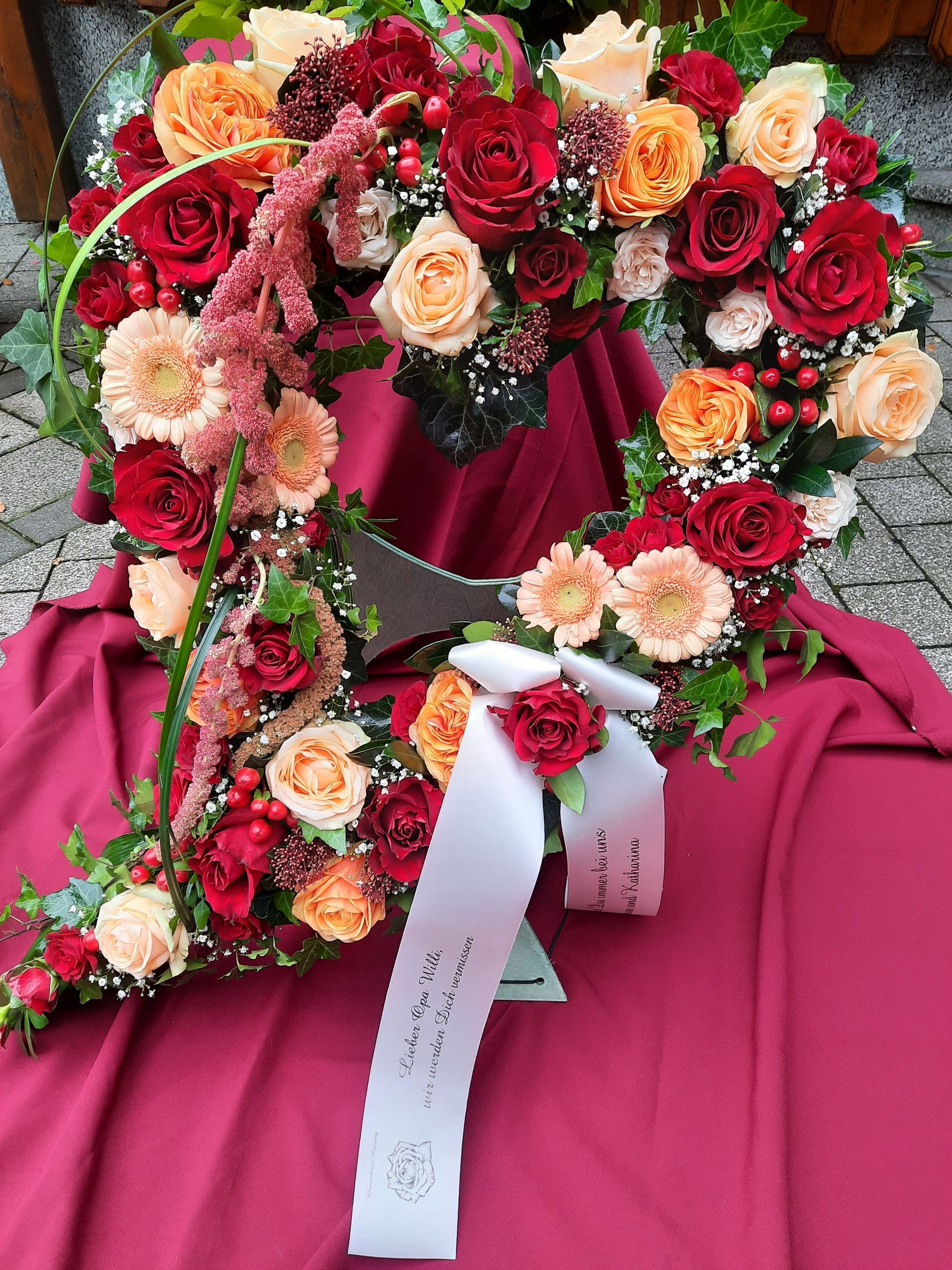 Herzförmiges Blumenarrangement in Rot- und Pfirsichtönen, mit weißem Band, auf einem burgunderfarbenen Tuch.