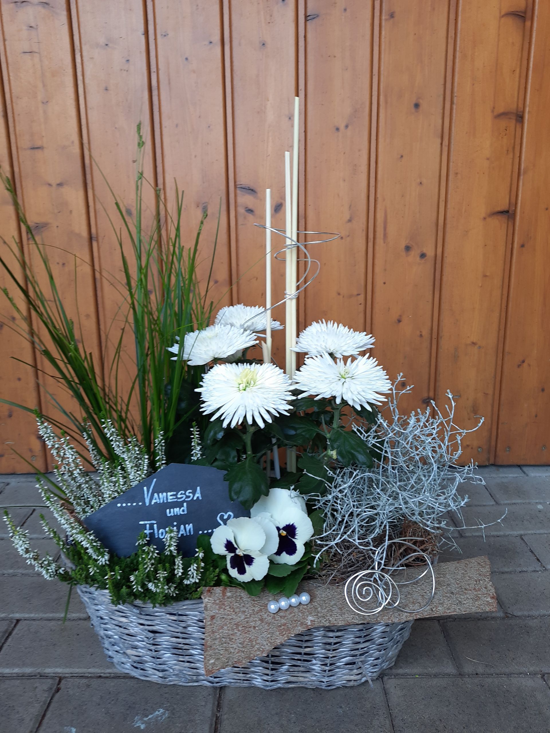 Blumenarrangement in einem geflochtenen Korb mit weißen Chrysanthemen, Stiefmütterchen und Dekoelementen.