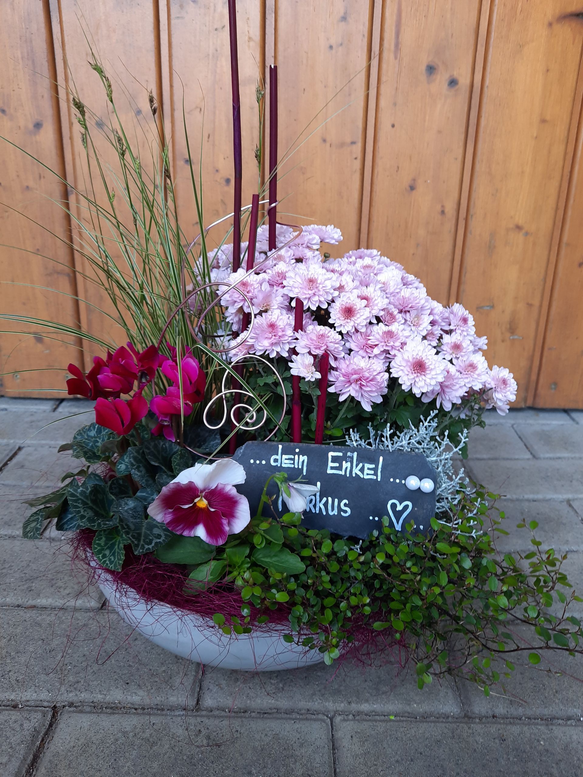 Ein Blumenarrangement in einer weißen Schale, darunter rosa Blumen, grünes Laub und ein handgeschriebenes Schild.