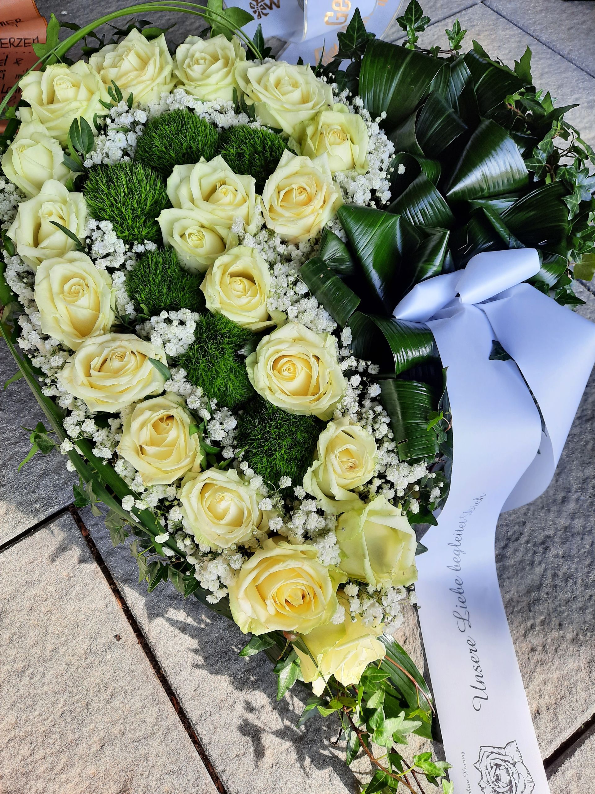 Trauergesteck aus weißen Rosen, Grünpflanzen und einem weißen Band auf einer Steinoberfläche.