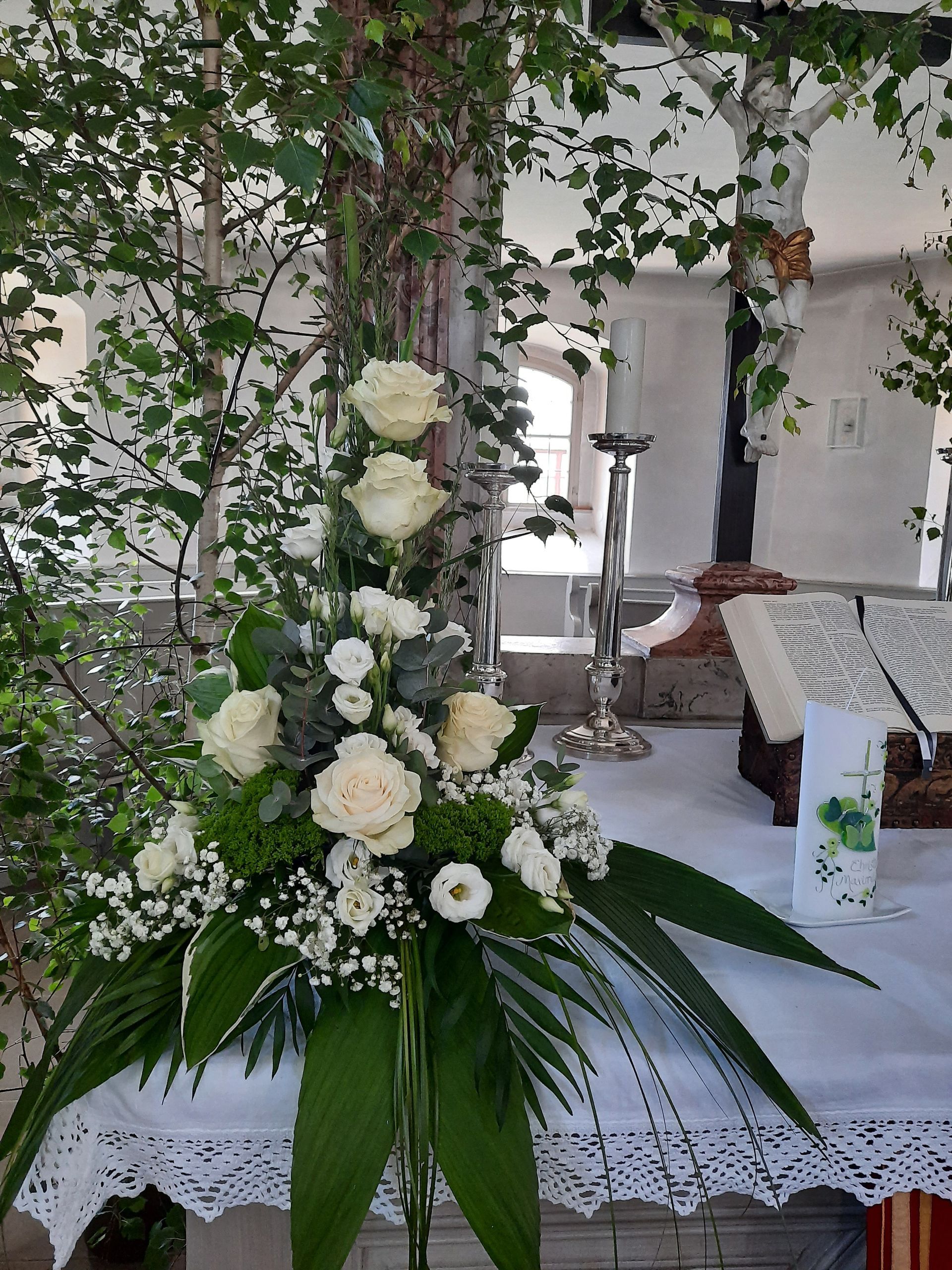 Weißes Blumenarrangement auf einem Tisch; weiße Rosen, Grünpflanzen und Kerzen, ein religiöser Rahmen.