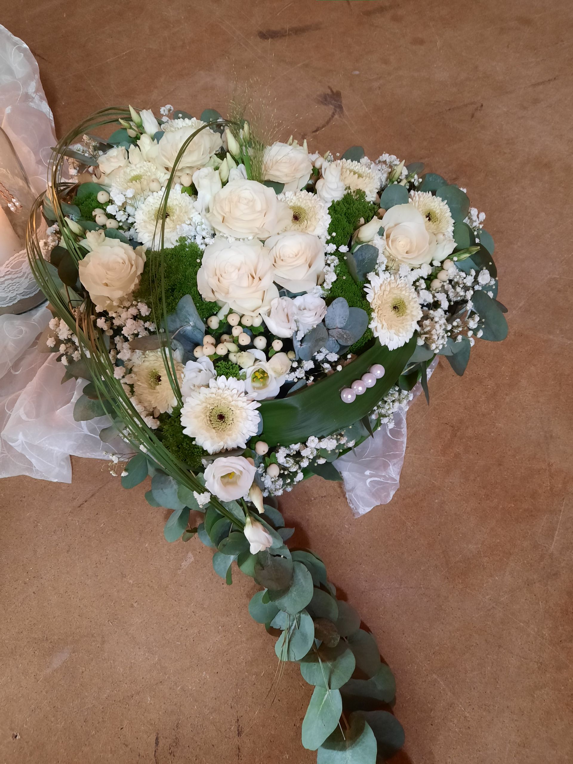 Herzförmiges Blumenarrangement mit weißen Rosen, Gänseblümchen und Grünpflanzen auf einer Holzoberfläche.