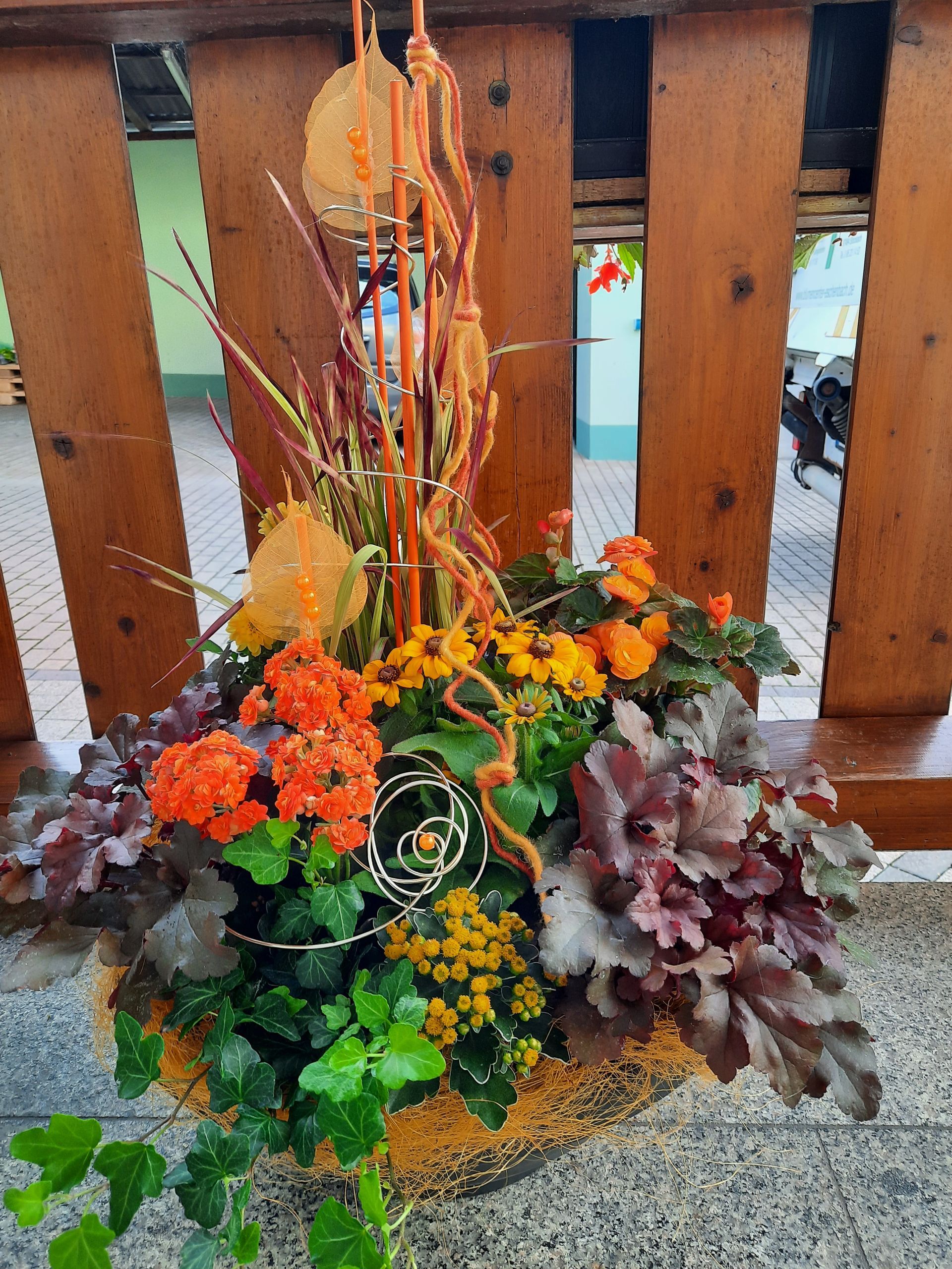 Ein Hängekorb voller bunter Herbstblumen, Blätter und dekorativer Elemente vor Holzbrettern.