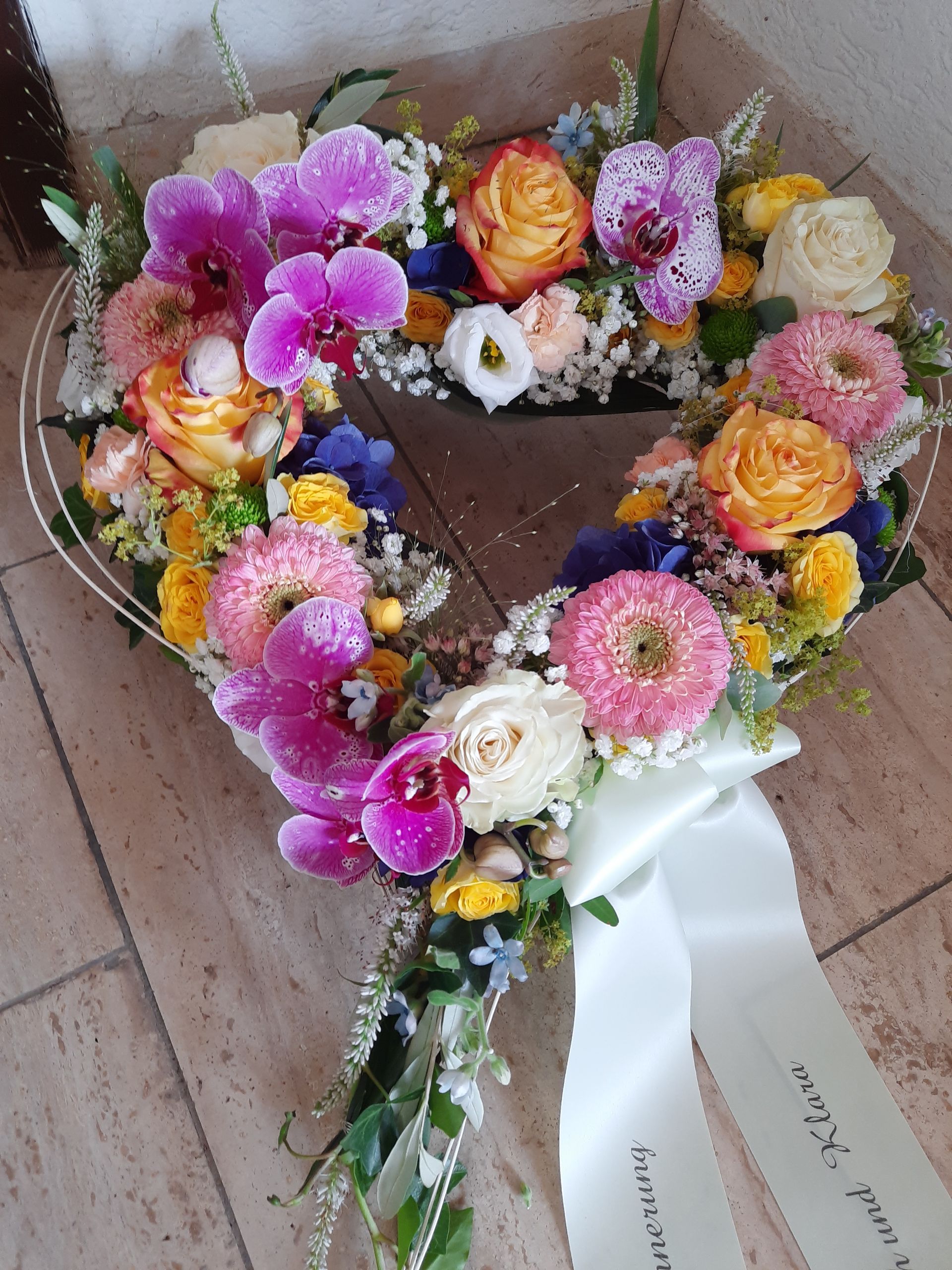 Herzförmiges Blumenarrangement mit rosa, orangefarbenen und weißen Blumen und einem weißen Band; auf einem Marmorboden ruhend.