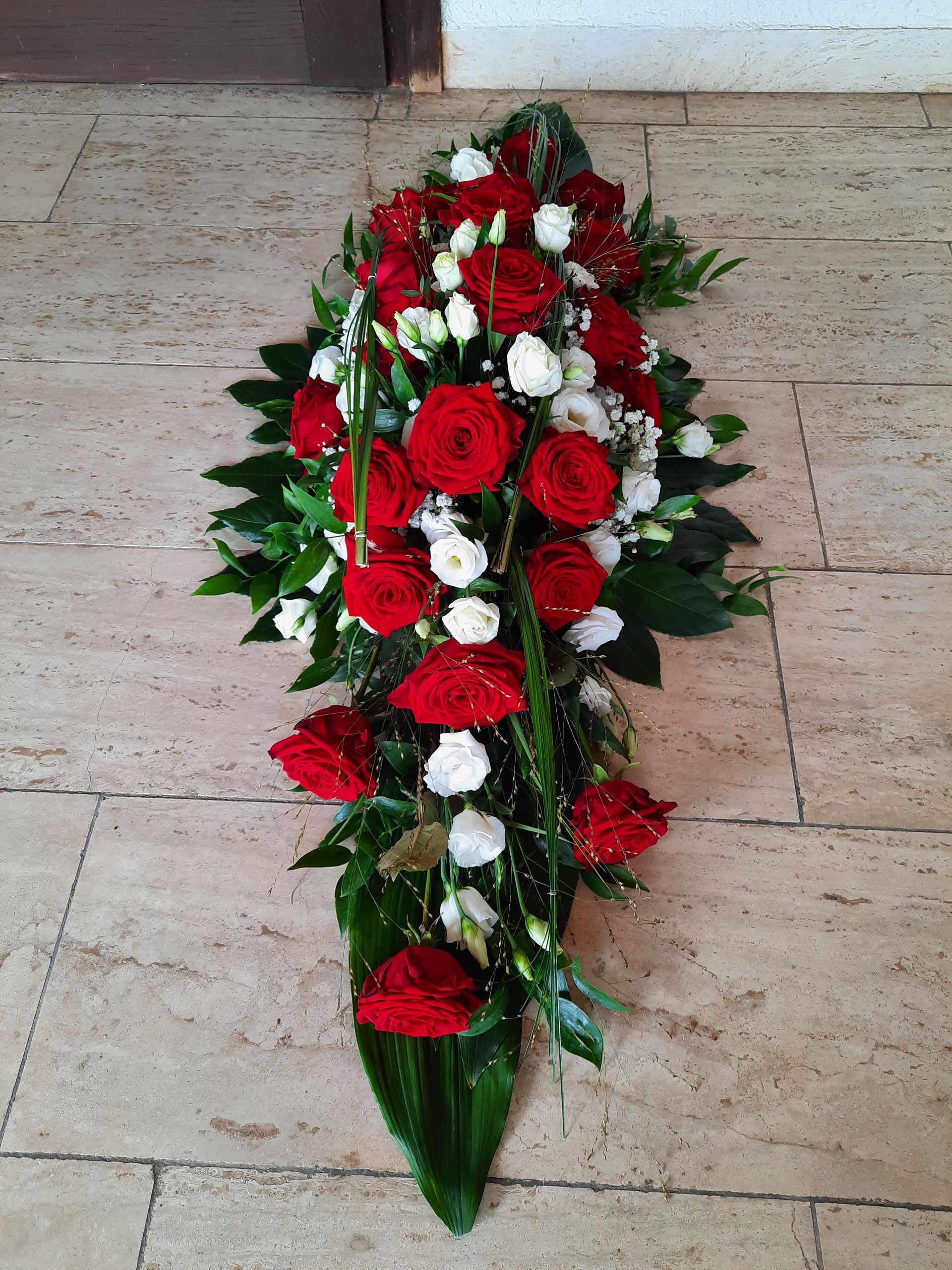 Rote und weiße Rosen sind in einem langen, tropfenförmigen Blumenarrangement angeordnet. Grüne Blätter rahmen die Blumen auf einem gefliesten Boden ein.
