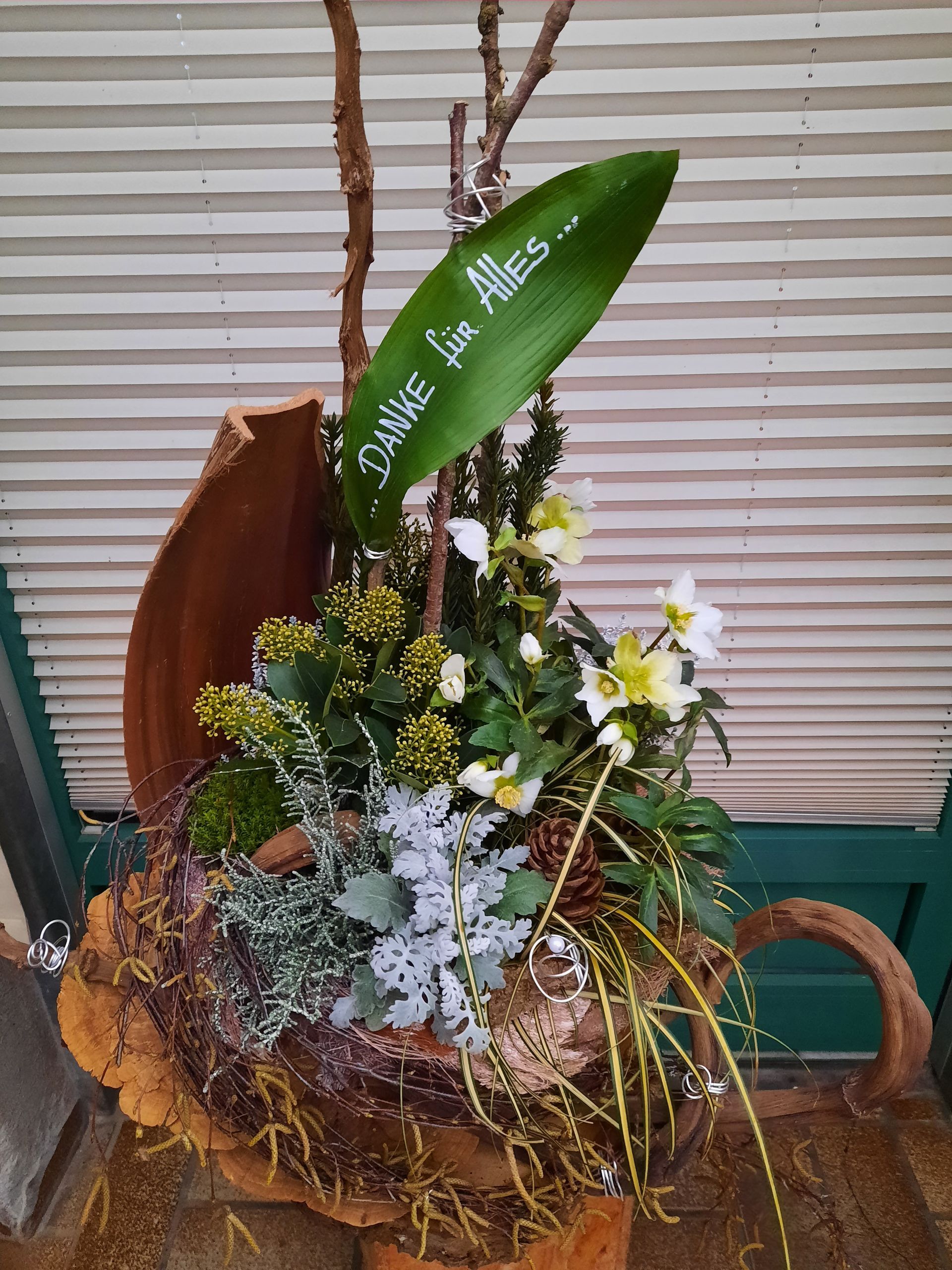 Blumenarrangement in einer geschnitzten Holztasse mit Blumen und Grünpflanzen. Grünes Blatt mit der Aufschrift „Danke für Alles“.