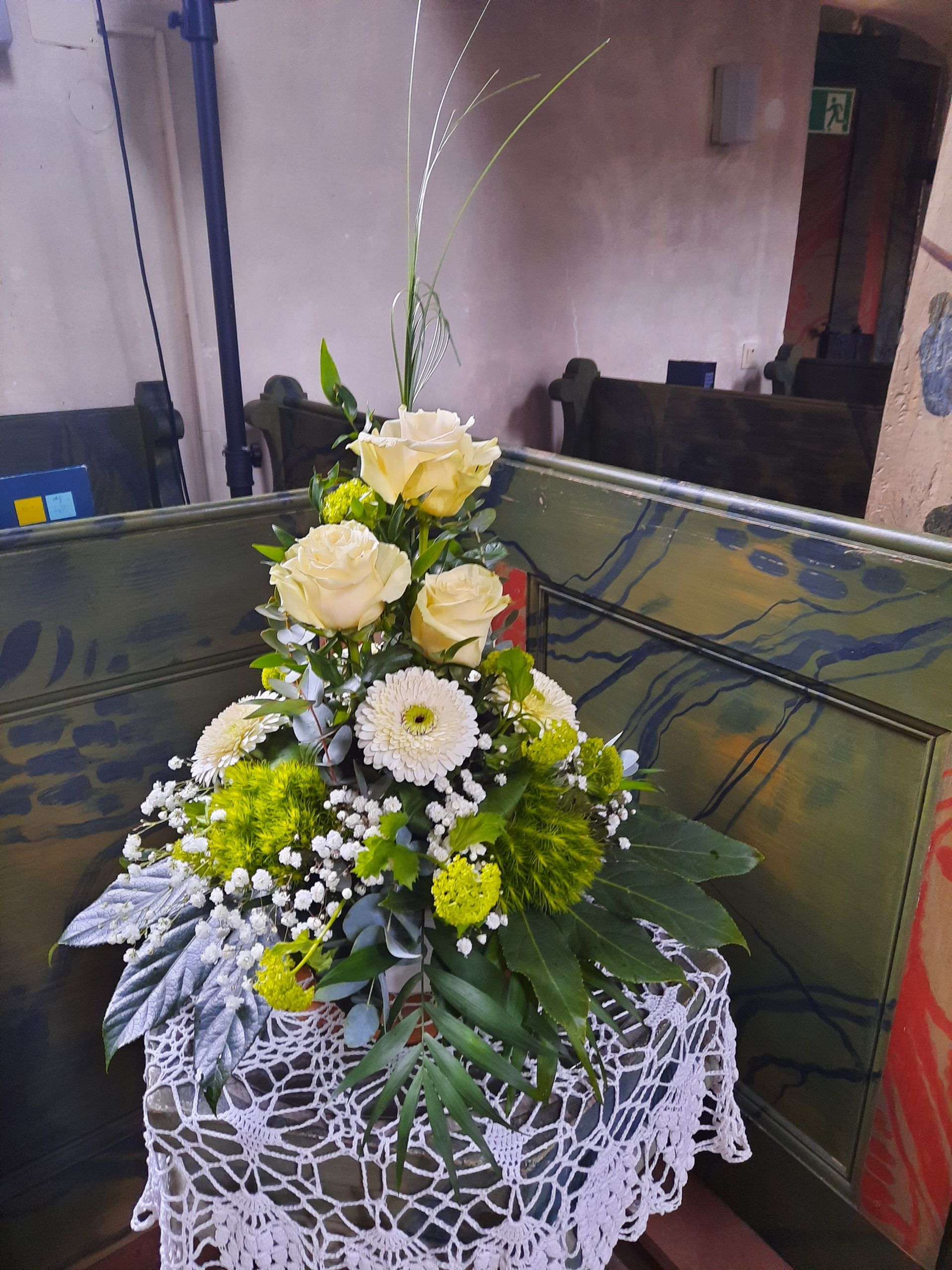 Blumenarrangement mit weißen Rosen, grünen Blüten und Blättern auf einer gehäkelten Deckchen in einer Kirche.