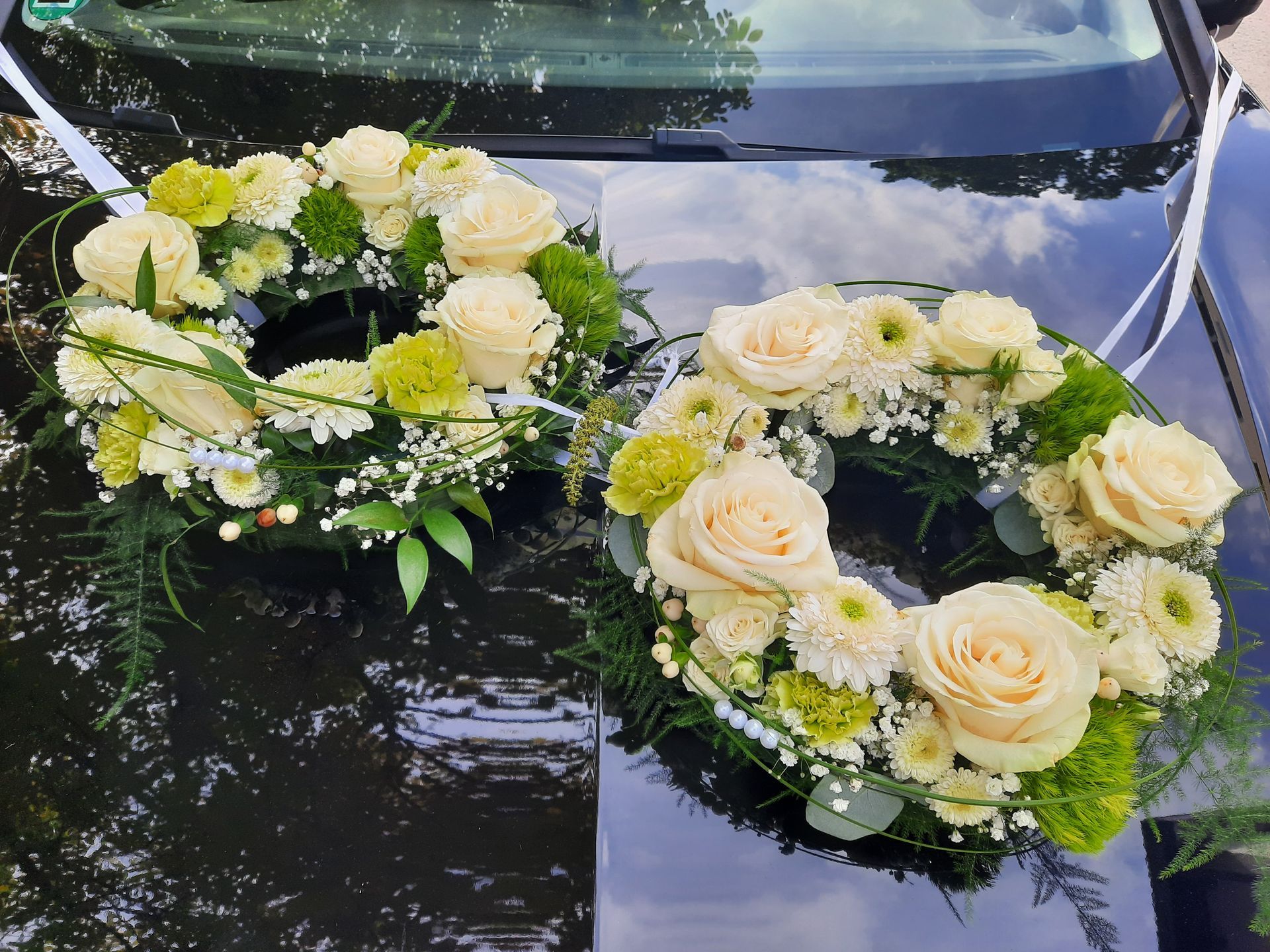 Zwei Blumenkränze aus cremefarbenen Rosen und grünen Akzenten zieren die Motorhaube eines dunklen Autos, möglicherweise für eine Hochzeit.