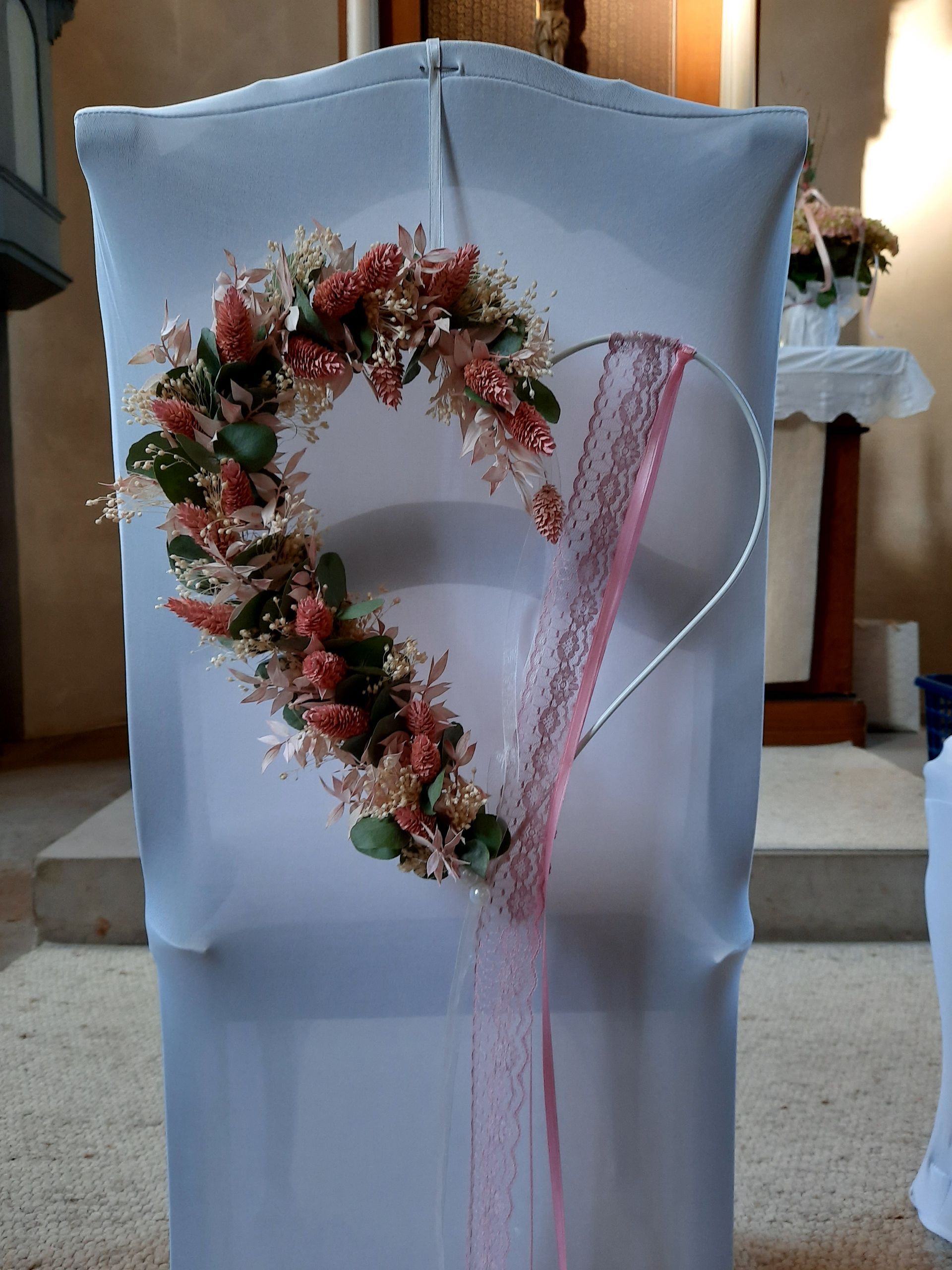Weiß bezogener Stuhl mit floralem Herzkranz und rosa Spitzenband in einer Kirche.