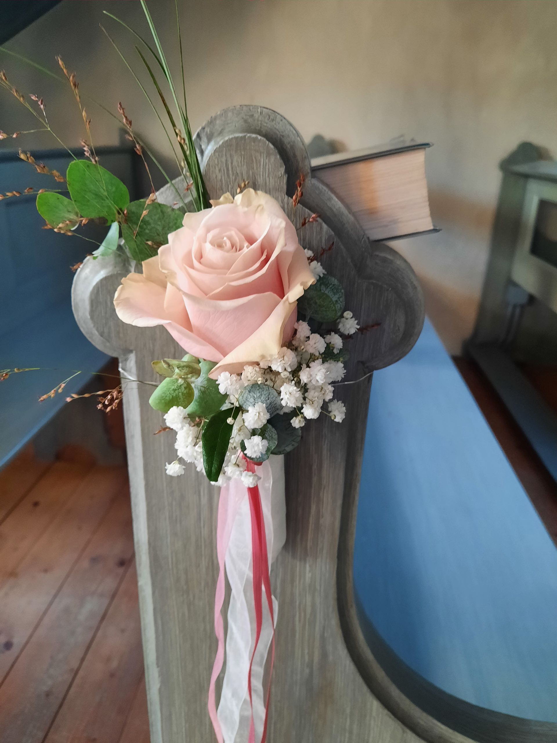 Blumenarrangement aus rosa Rosen auf einer hellblauen Kirchenbank.