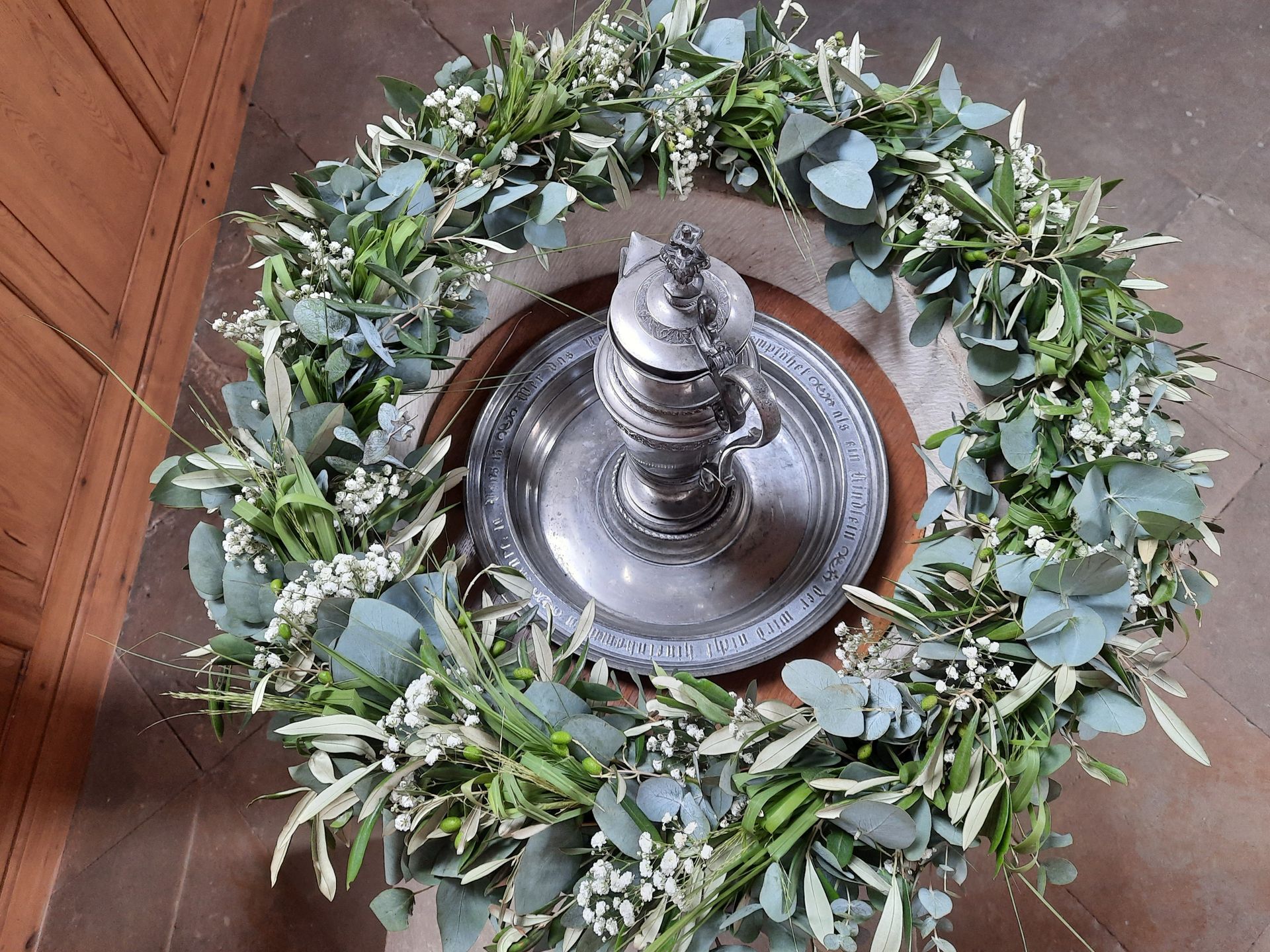 Silberne Kanne auf Tablett, umgeben von einem grün-weißen Blumenkranz, auf einer Holzoberfläche platziert.