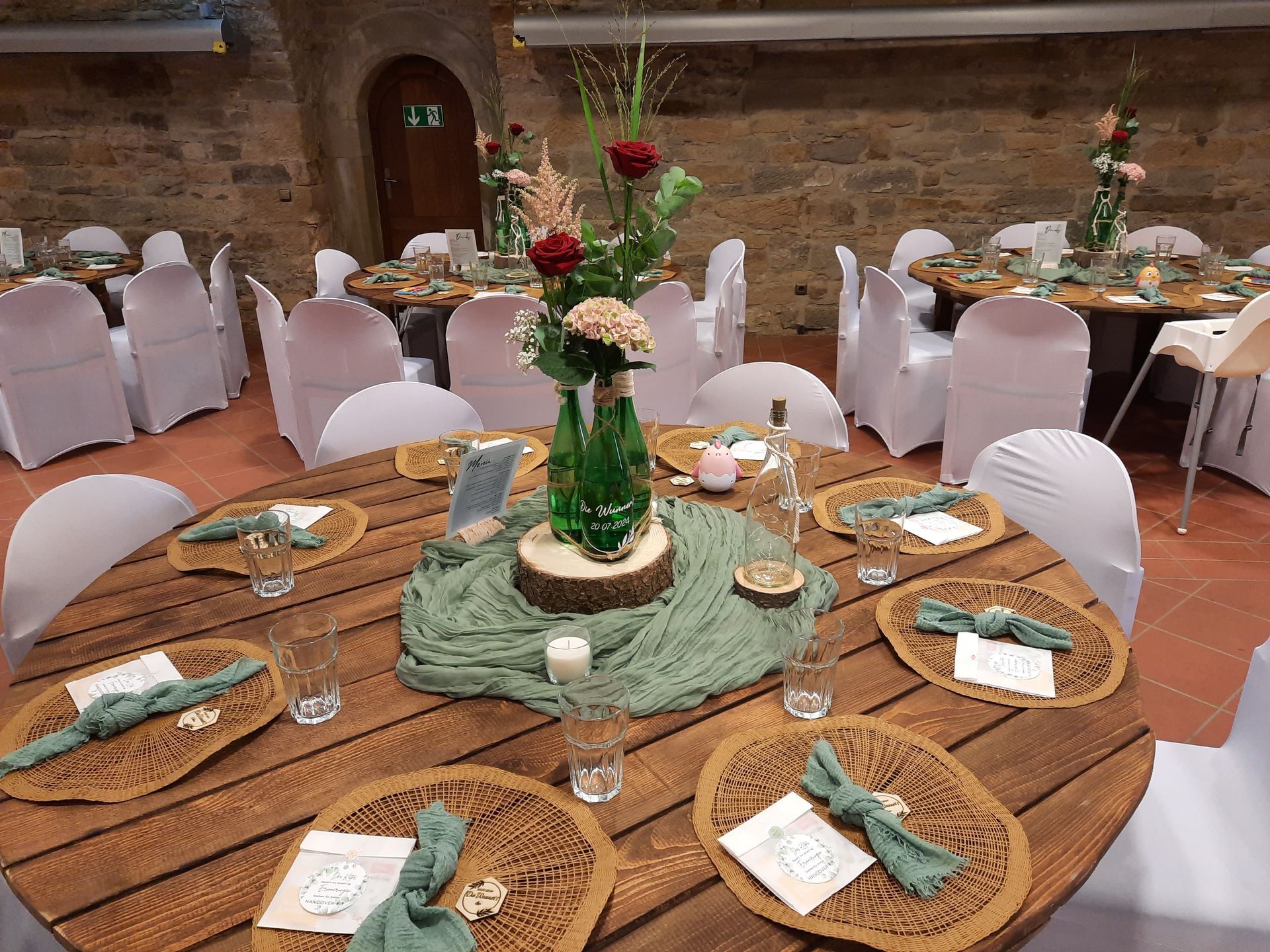 Tische für den Hochzeitsempfang mit Blumengestecken, Tischsets aus Holz und weißen Stuhlhussen in einem rustikalen Saal.