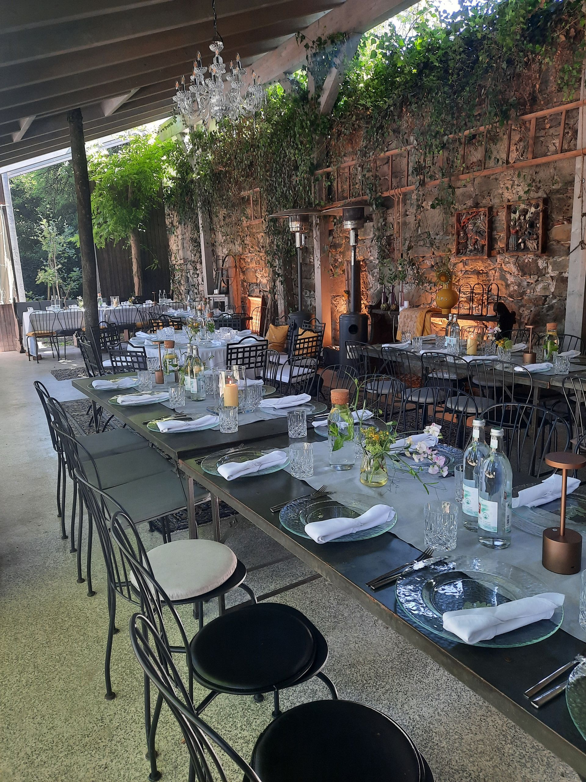 Essbereich im Freien: lange Tische, Stühle und Gedecke vor einer mit Laub bedeckten Backsteinmauer.