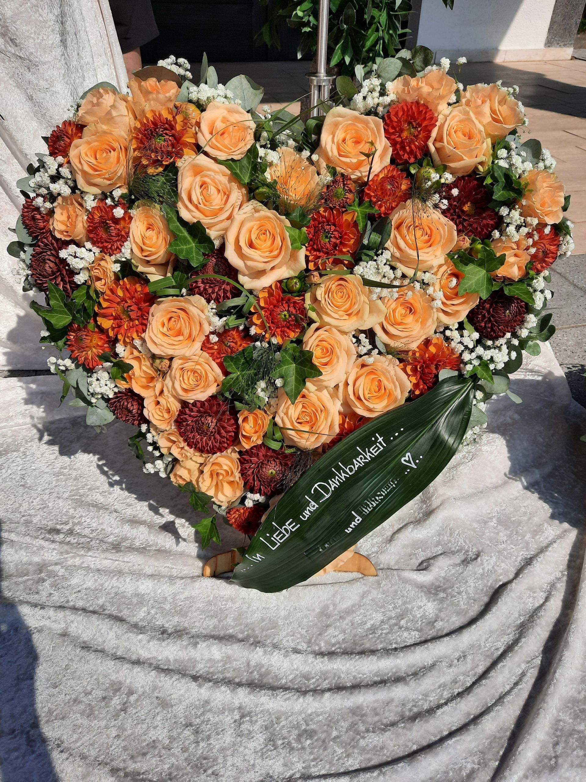 Herzförmiges Blumenarrangement mit pfirsich- und orangefarbenen Rosen, dunkelroten Blüten und einer grünen Aufschrift auf weißem Untergrund.