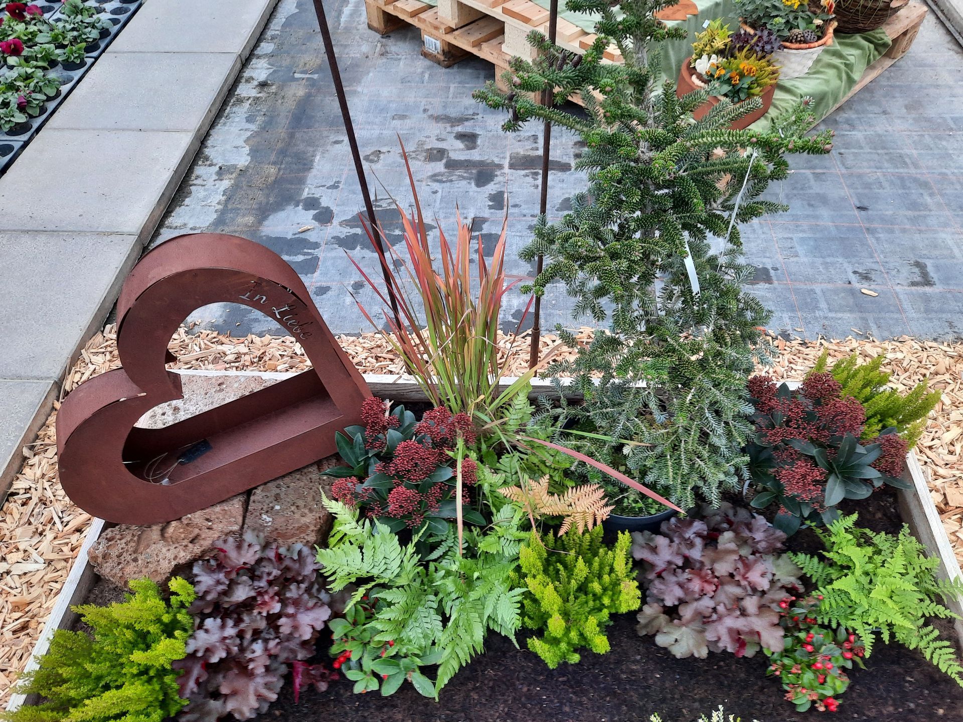 Eine Gartenausstellung mit einer rostigen Herzskulptur, verschiedenen Pflanzen und Holzspänen.