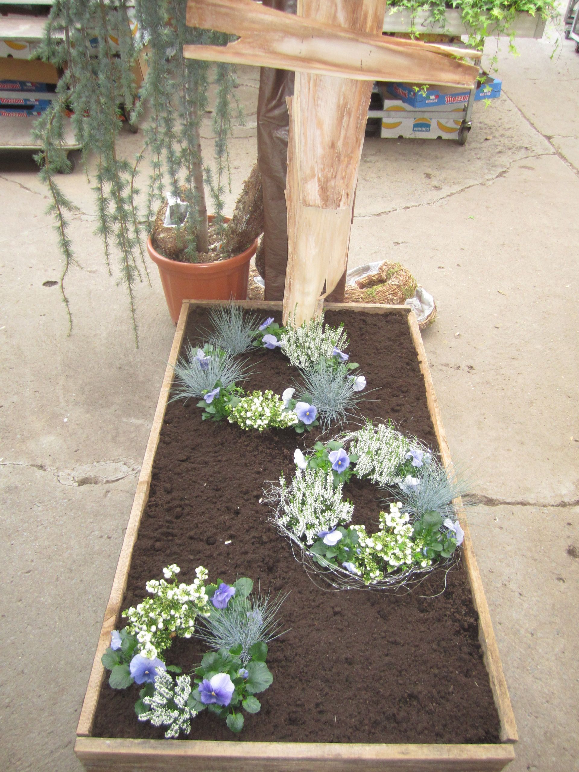 Holzpflanzgefäß mit drei Blumenkränzen in dunkler Erde, neben einem Holzkreuz.