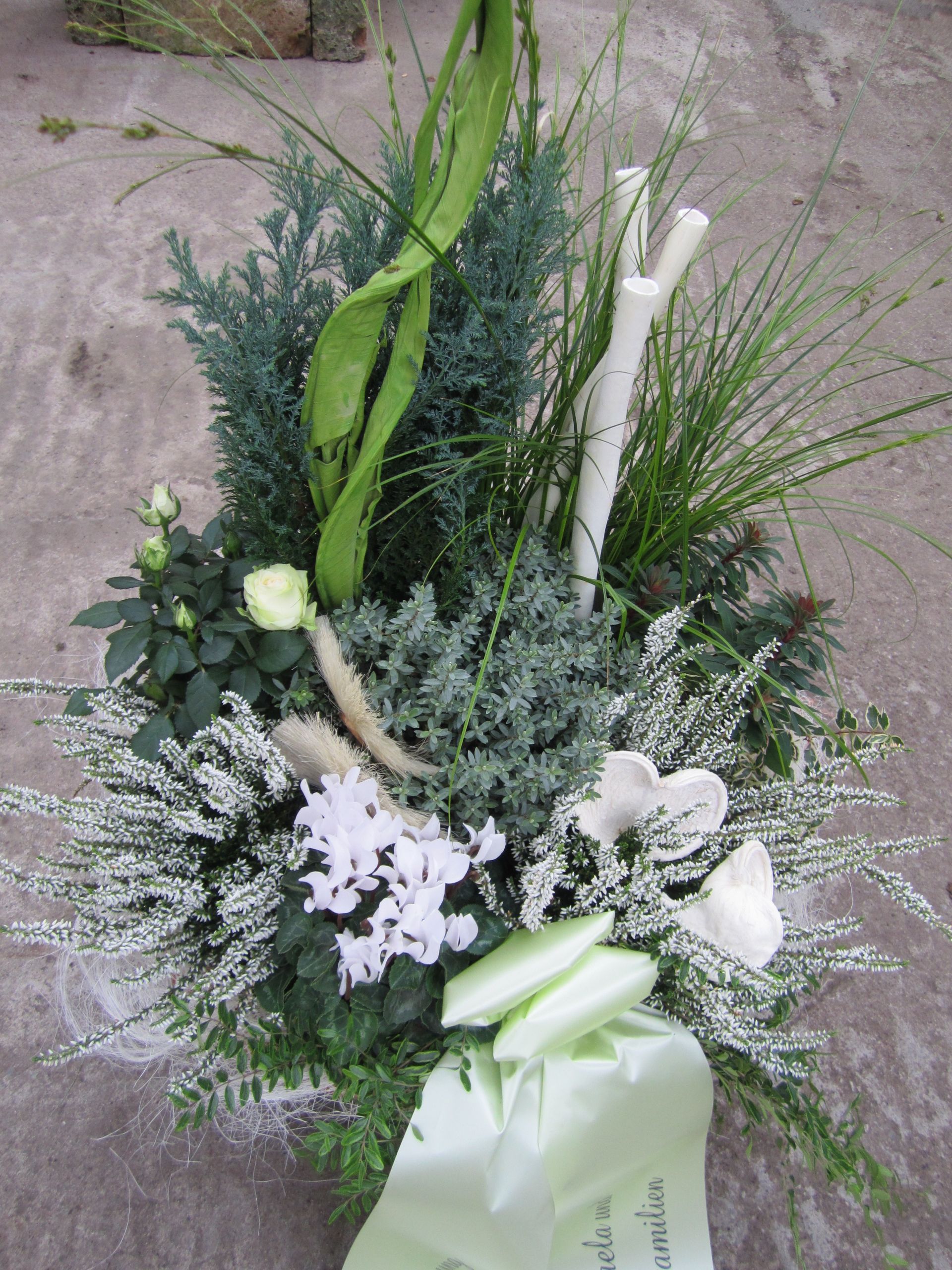 Trauergesteck mit weißen Blumen, blaugrünem Laub, einem hellgrünen Band und einem weißen Zweig.