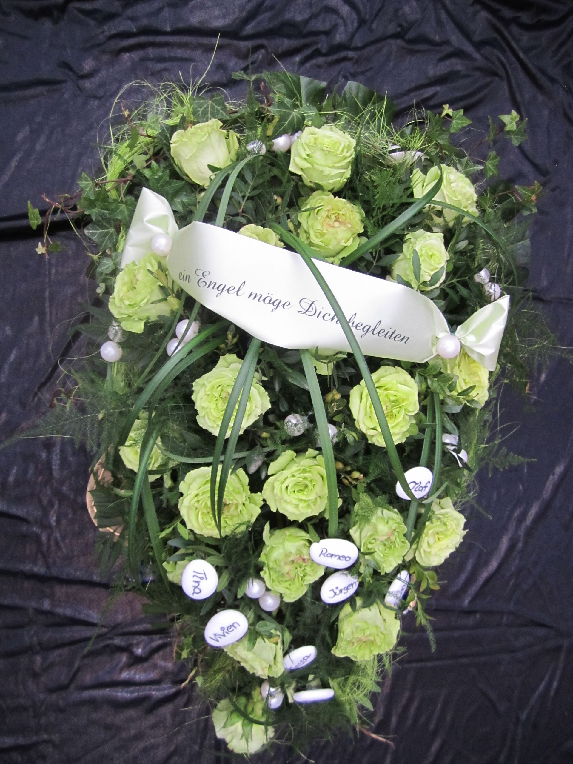 Grünes Blumenarrangement mit Schleife und weißen Namensschildern.