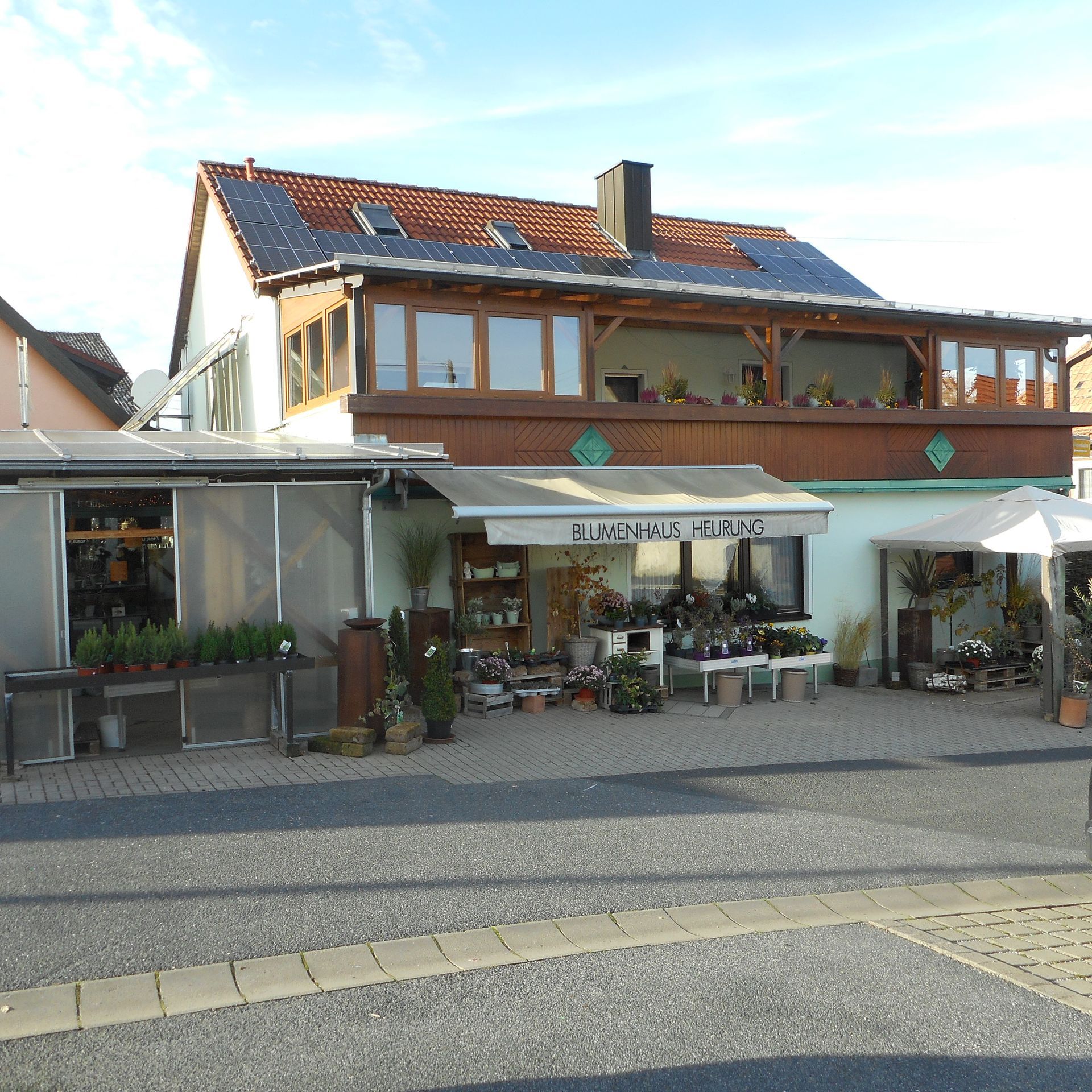 Außenansicht eines Blumenladens mit ausgestellten Pflanzen, brauner Markise, Balkon im zweiten Stock und Solarmodulen auf dem Dach.