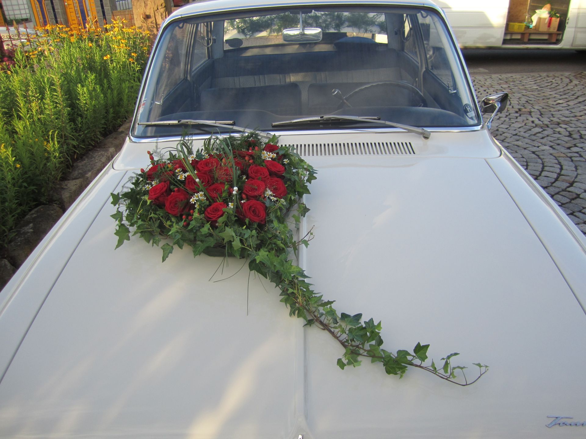 Cremefarbene Motorhaube eines Oldtimers, dekoriert mit roten Rosen und grünem Efeu; im Freien.